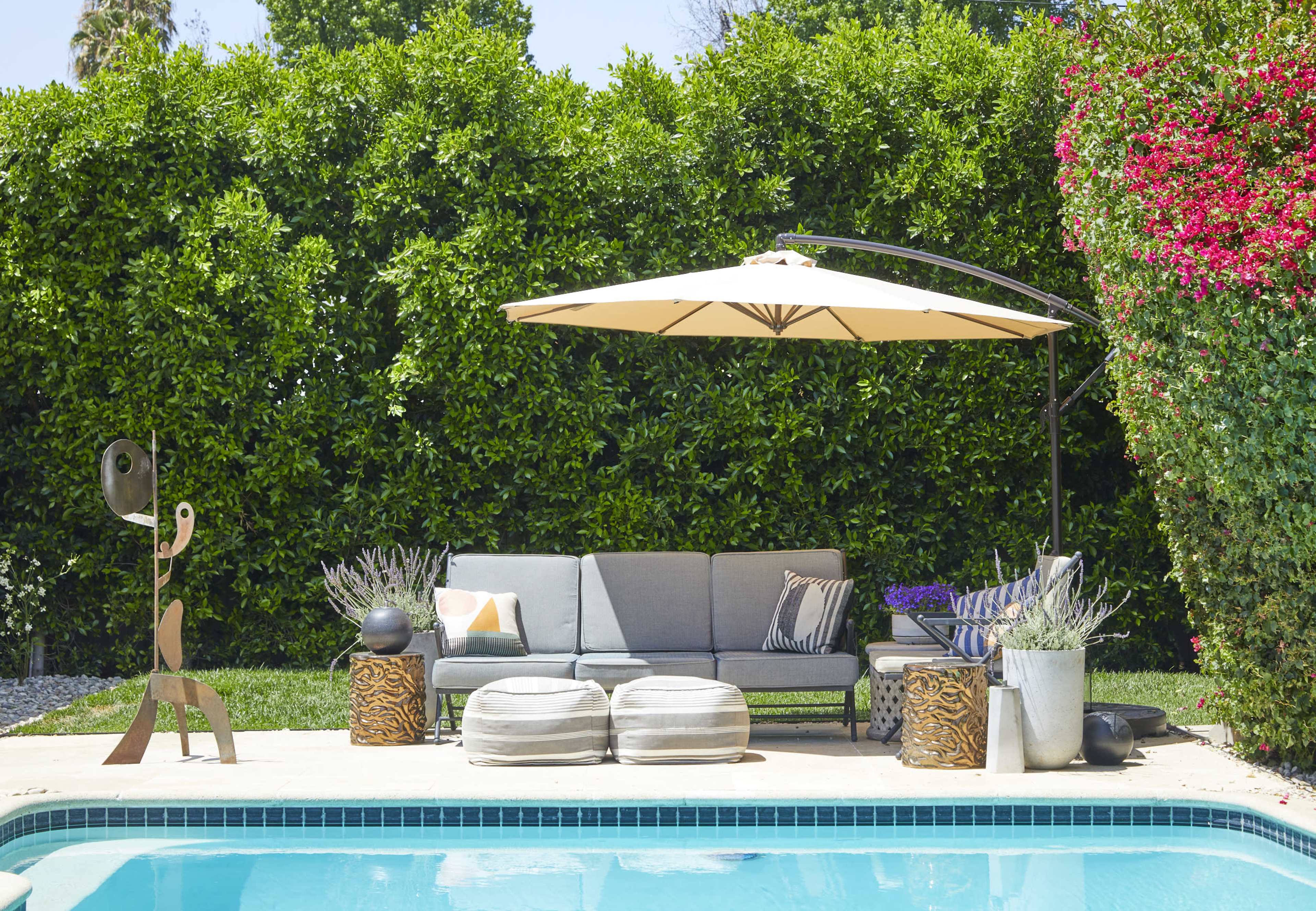 A modern outdoor seating area with a gray couch, large umbrella, decorative plants, and a pool in the foreground.