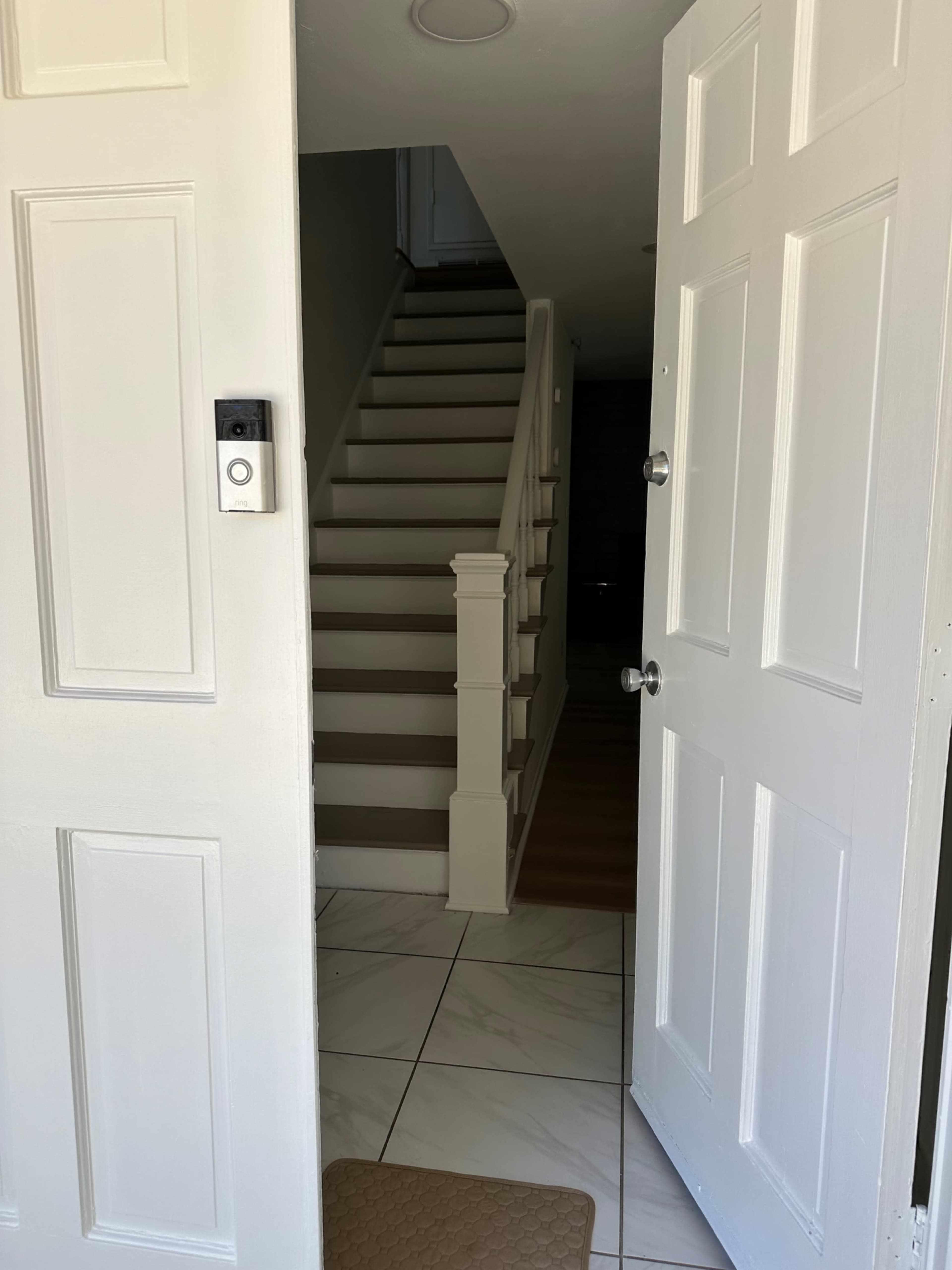 The image shows an open front door leading to a staircase with a white railing, inside a house.