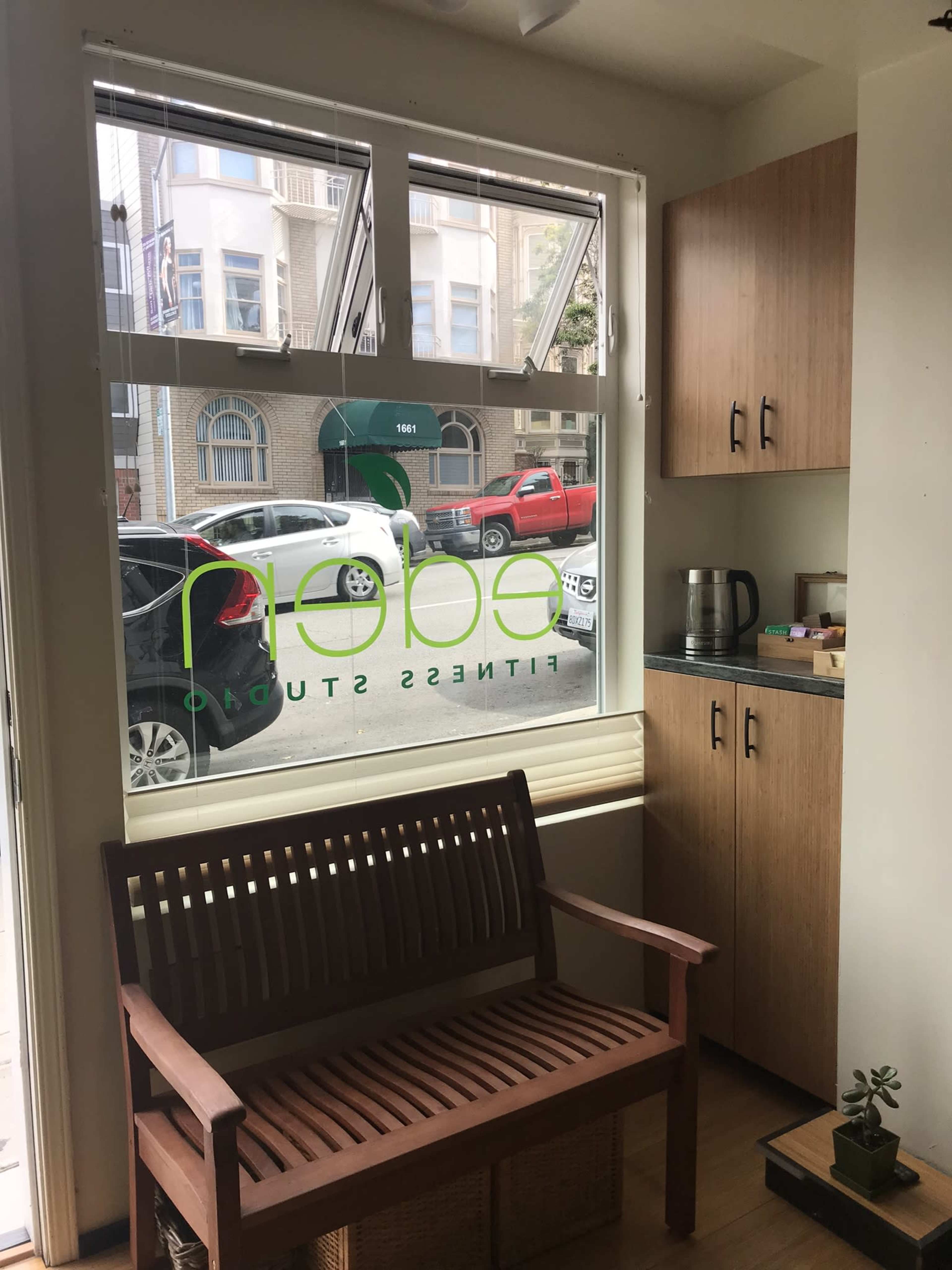 A fitness studio entrance features a wooden bench beside a kitchenette, with a large window displaying the studio's name and a view of the street outside.