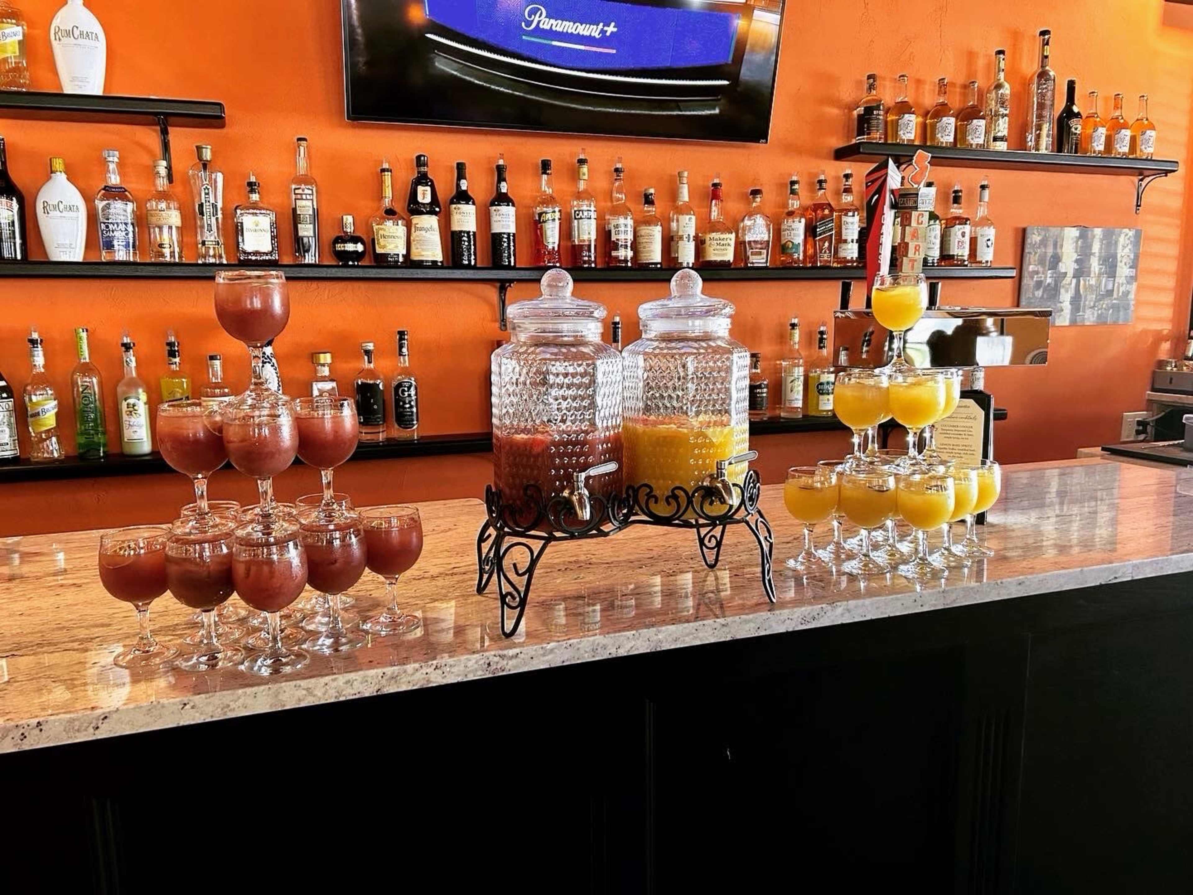 The image shows a bar setup with two beverage dispensers filled with different juices, surrounded by glassware and shelves of various alcoholic bottles on an orange wall.