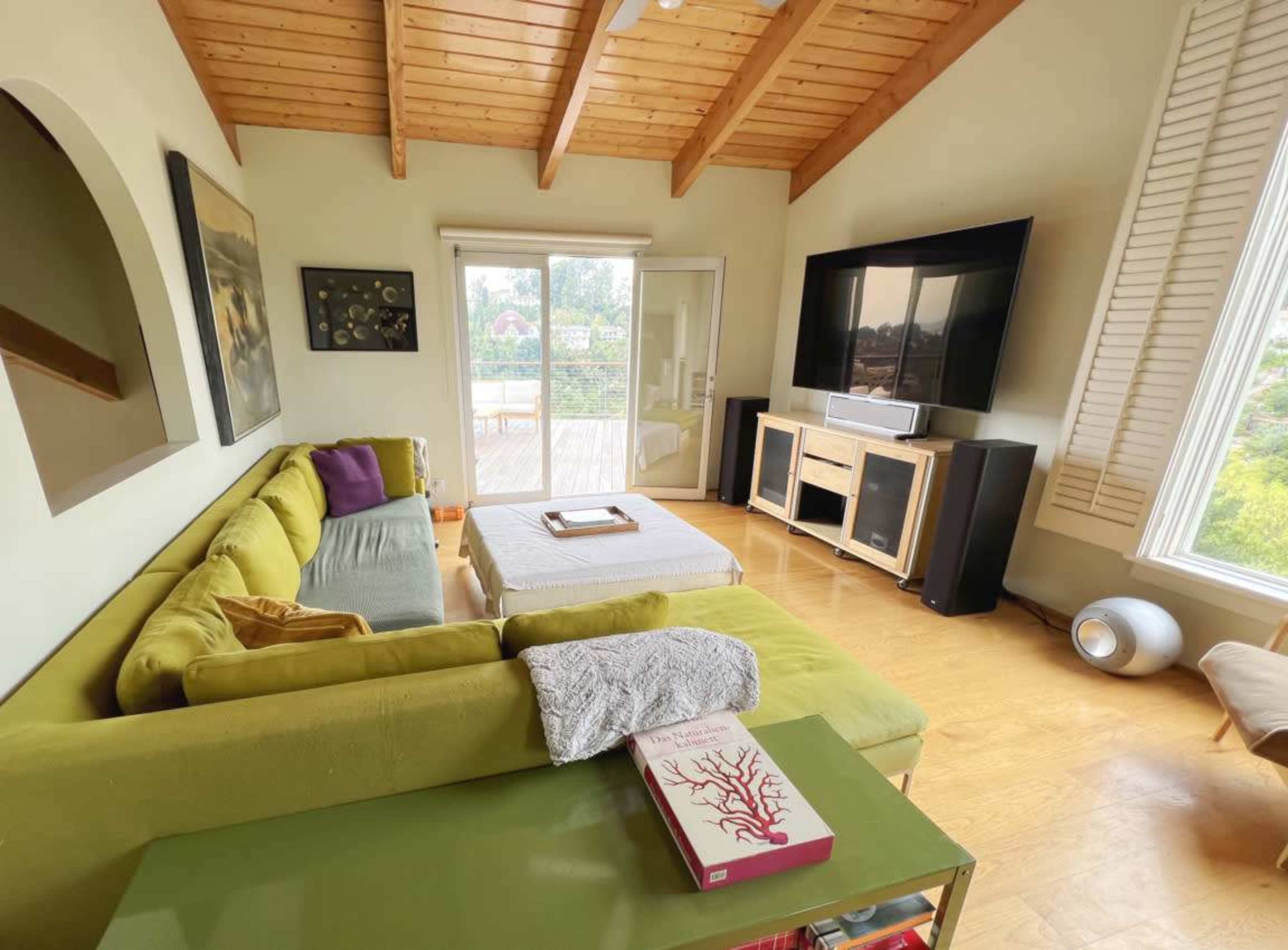 A modern living room features a green sectional sofa, a coffee table, a television mounted on the wall, and large windows with outdoor views.