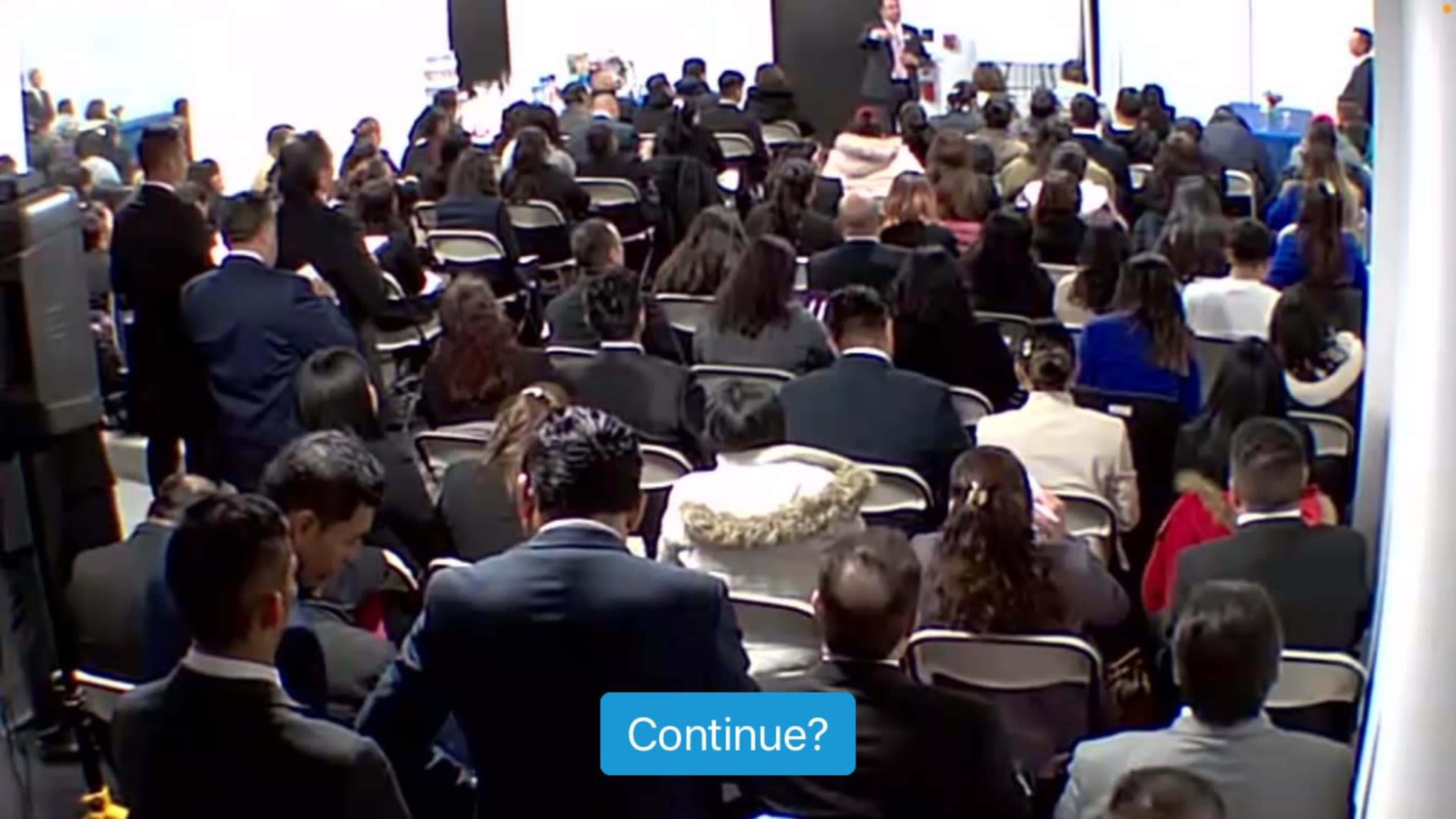 A large audience is seated in rows, attentively listening to a speaker at the front of the room.