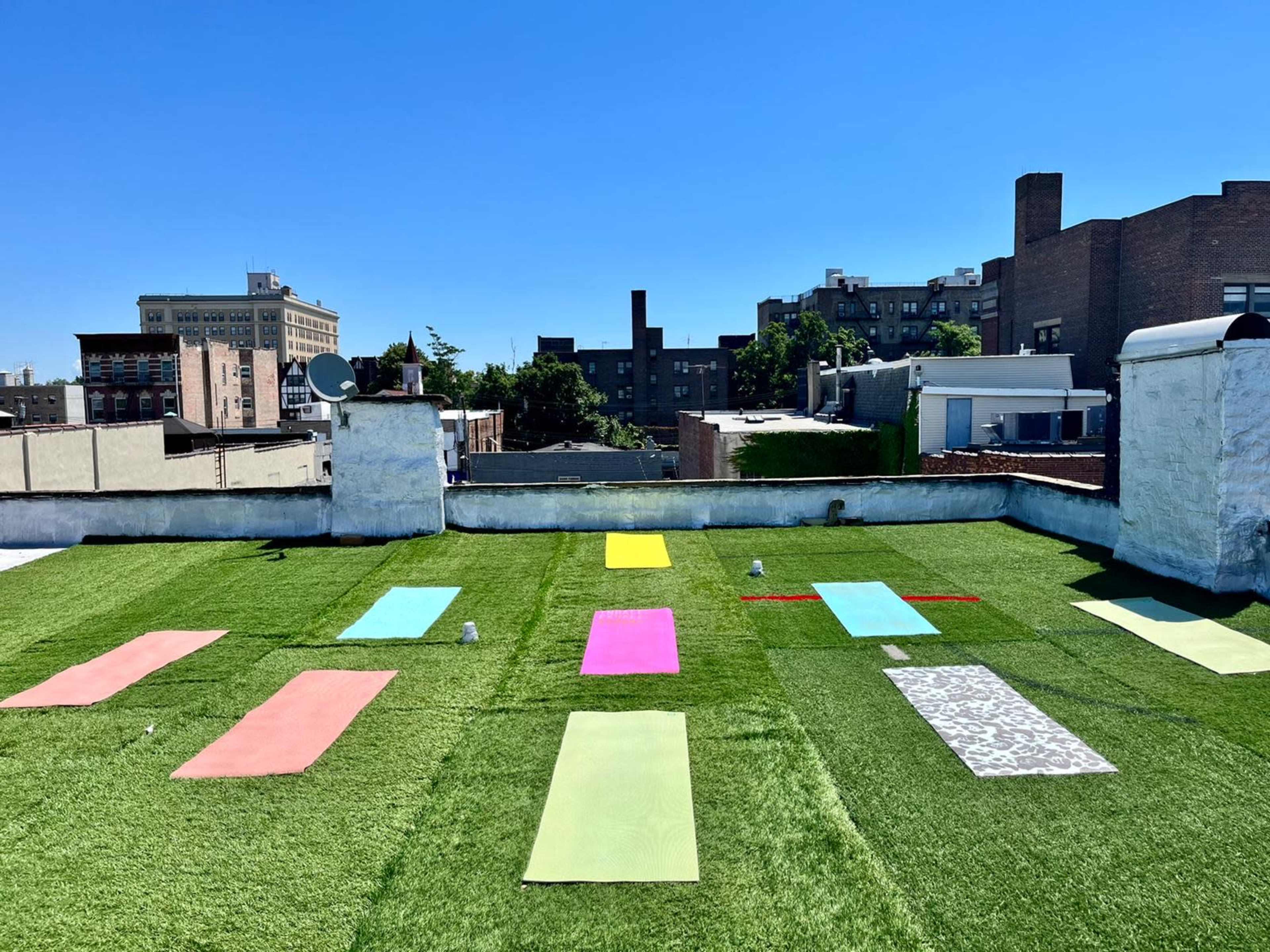 The image shows a rooftop yoga setup with several colorful mats arranged on green artificial grass under a clear blue sky.