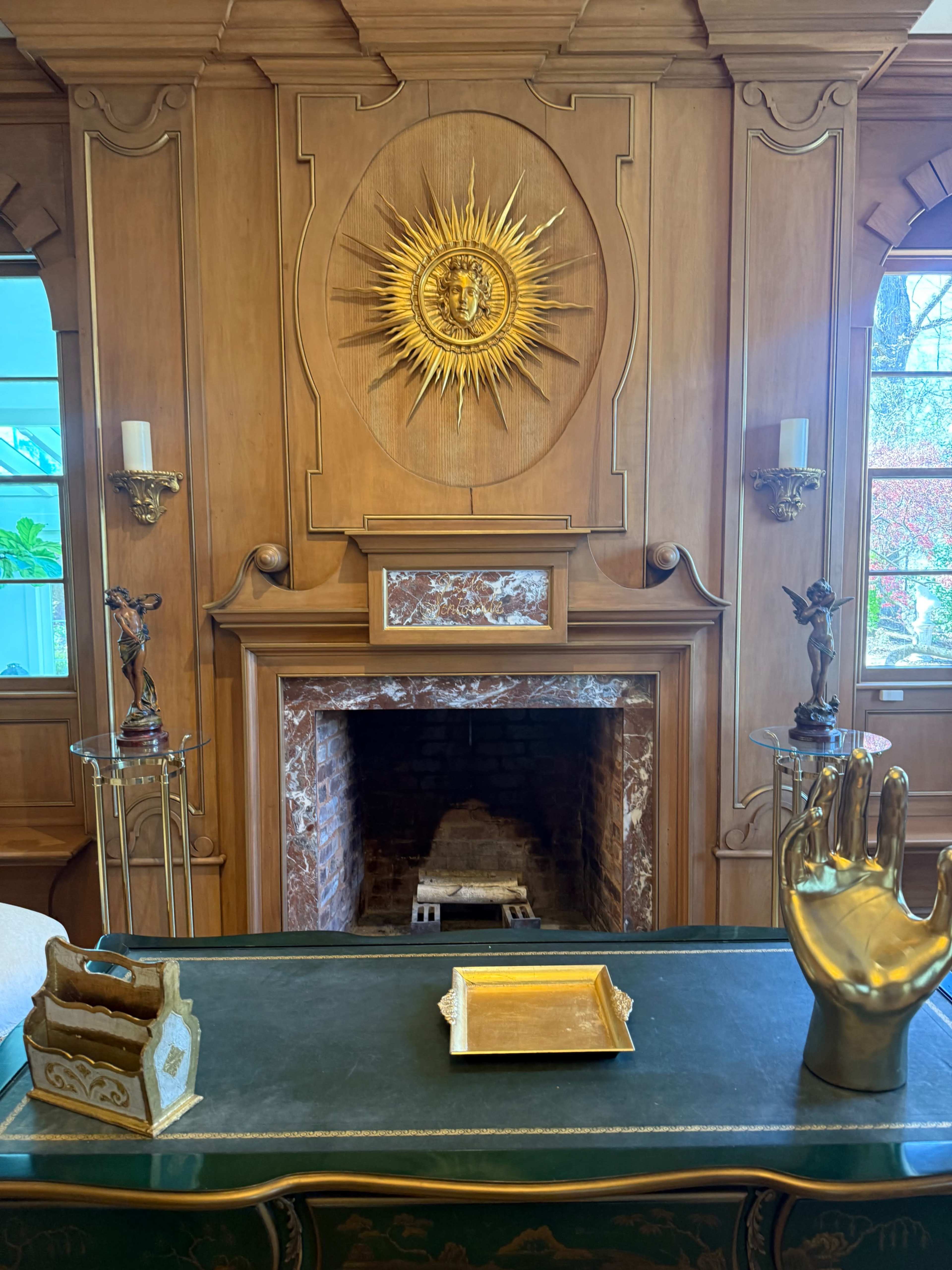 The image shows a wooden-paneled room featuring a decorative sunburst medallion above a fireplace, with a hand sculpture and a small tray on a green table in the foreground.