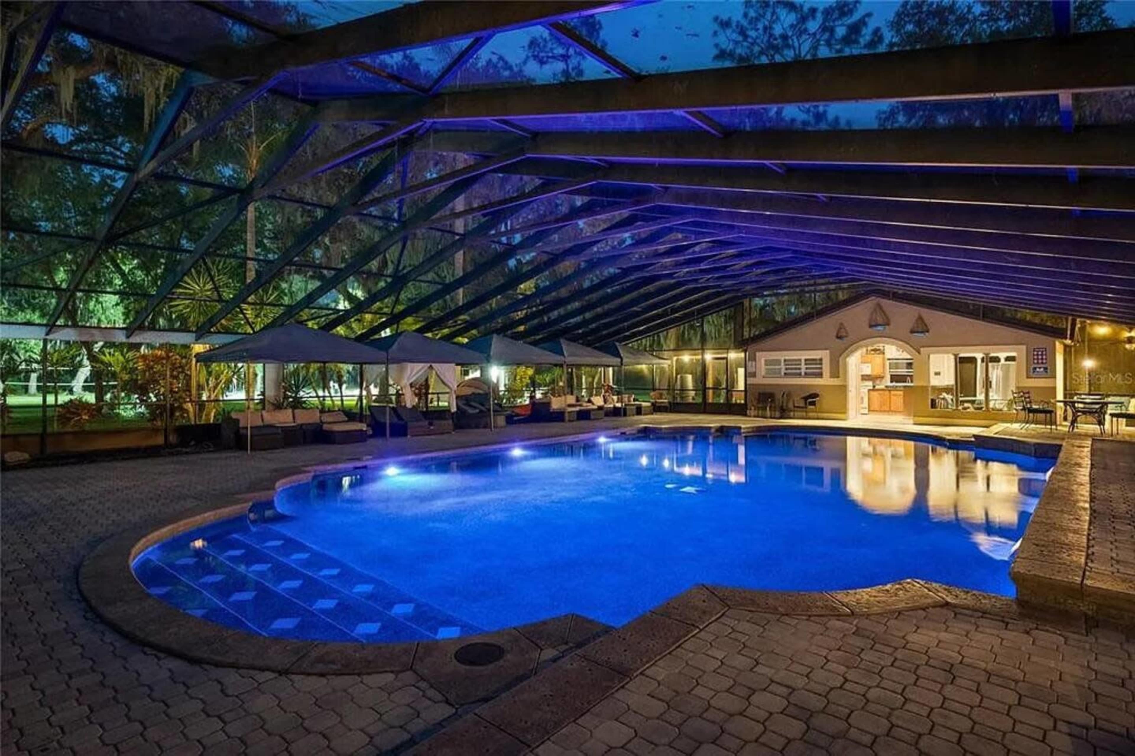 The image shows a spacious, illuminated swimming pool area enclosed by a glass structure, with lounge seating and a house in the background.