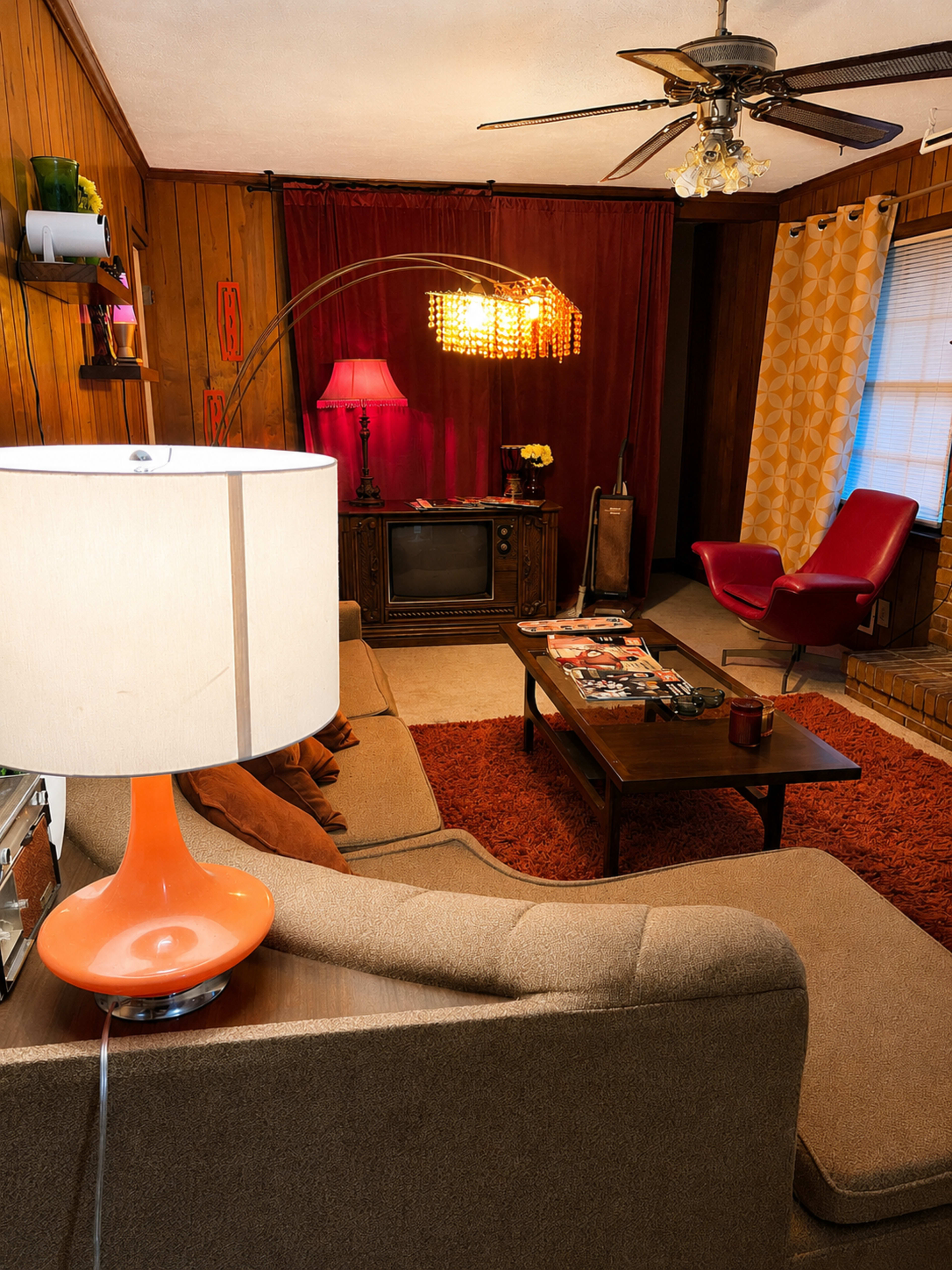 The image shows a vintage-inspired living room with a sofa, coffee table, lamp, and a television, surrounded by wooden walls and decorative curtains.