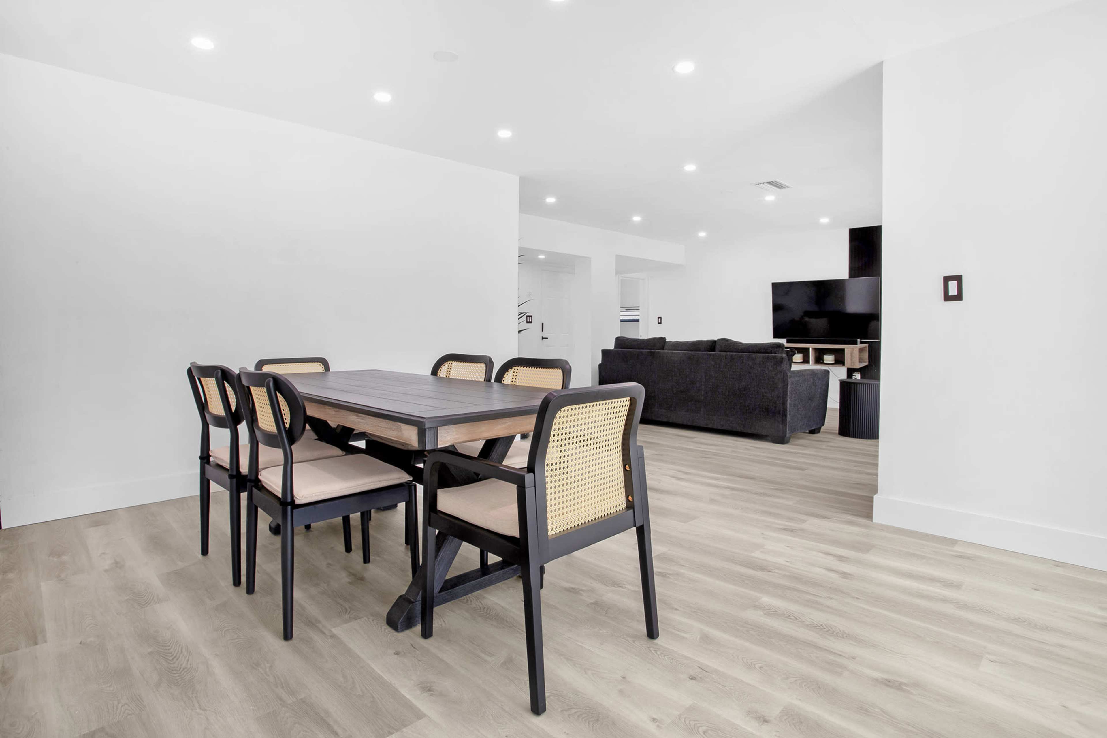 The image shows a modern dining area with a wooden table and six chairs, adjacent to a spacious living room featuring a dark sofa and a television.
