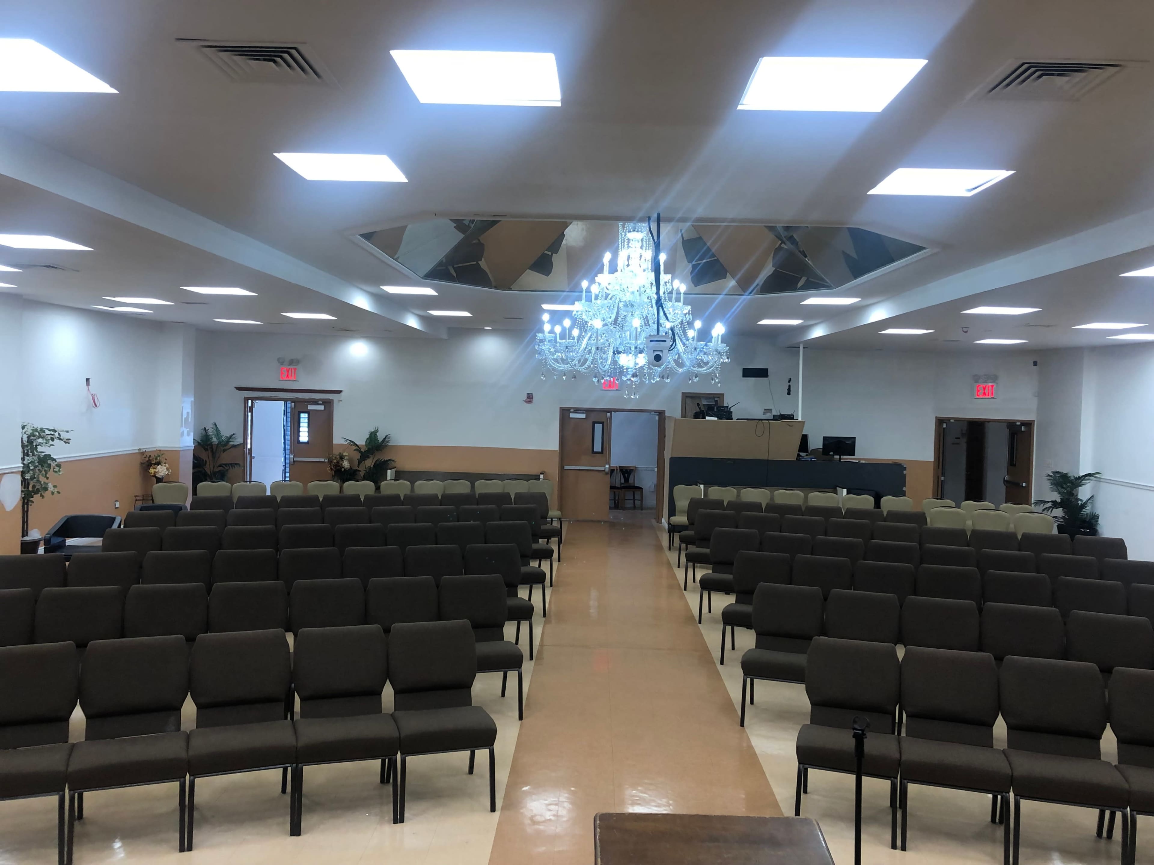 A large, well-lit empty hall with rows of brown chairs facing a stage area under a chandelier.