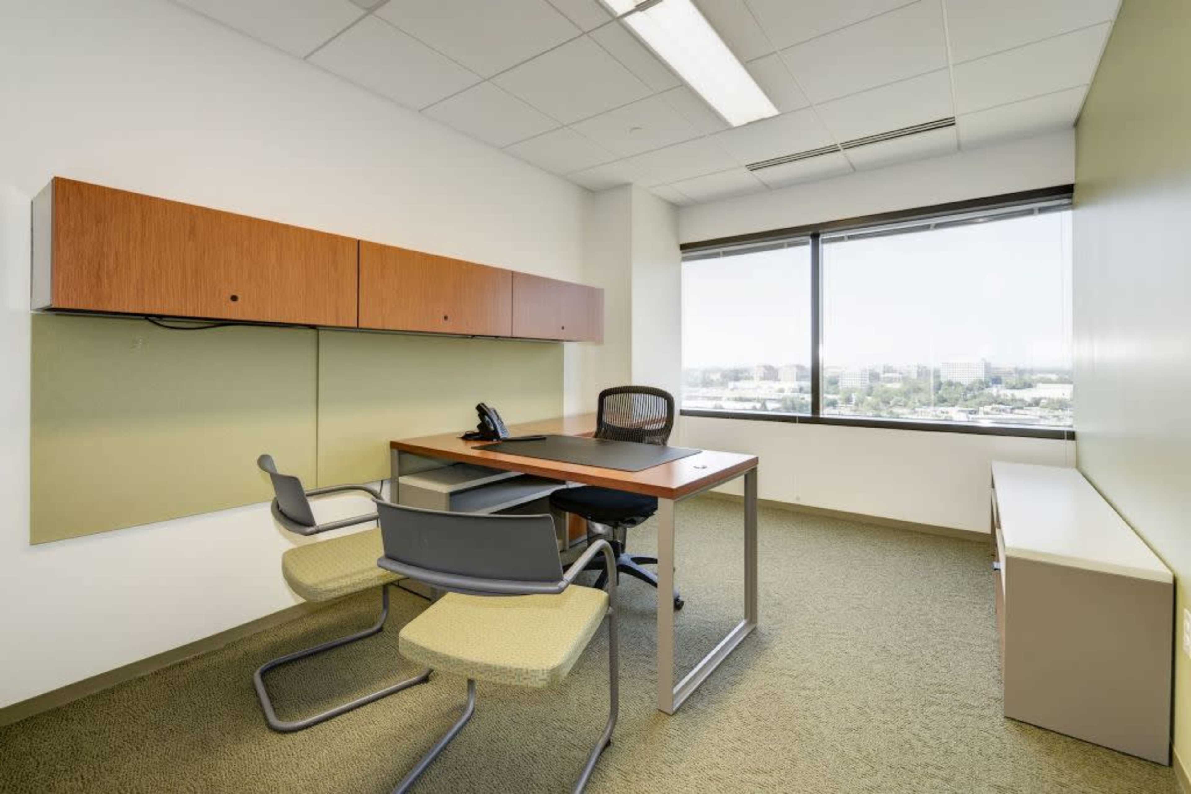 The image shows a minimalist office space featuring a desk with a chair, a telephone, and a wall-mounted storage unit beside a large window.