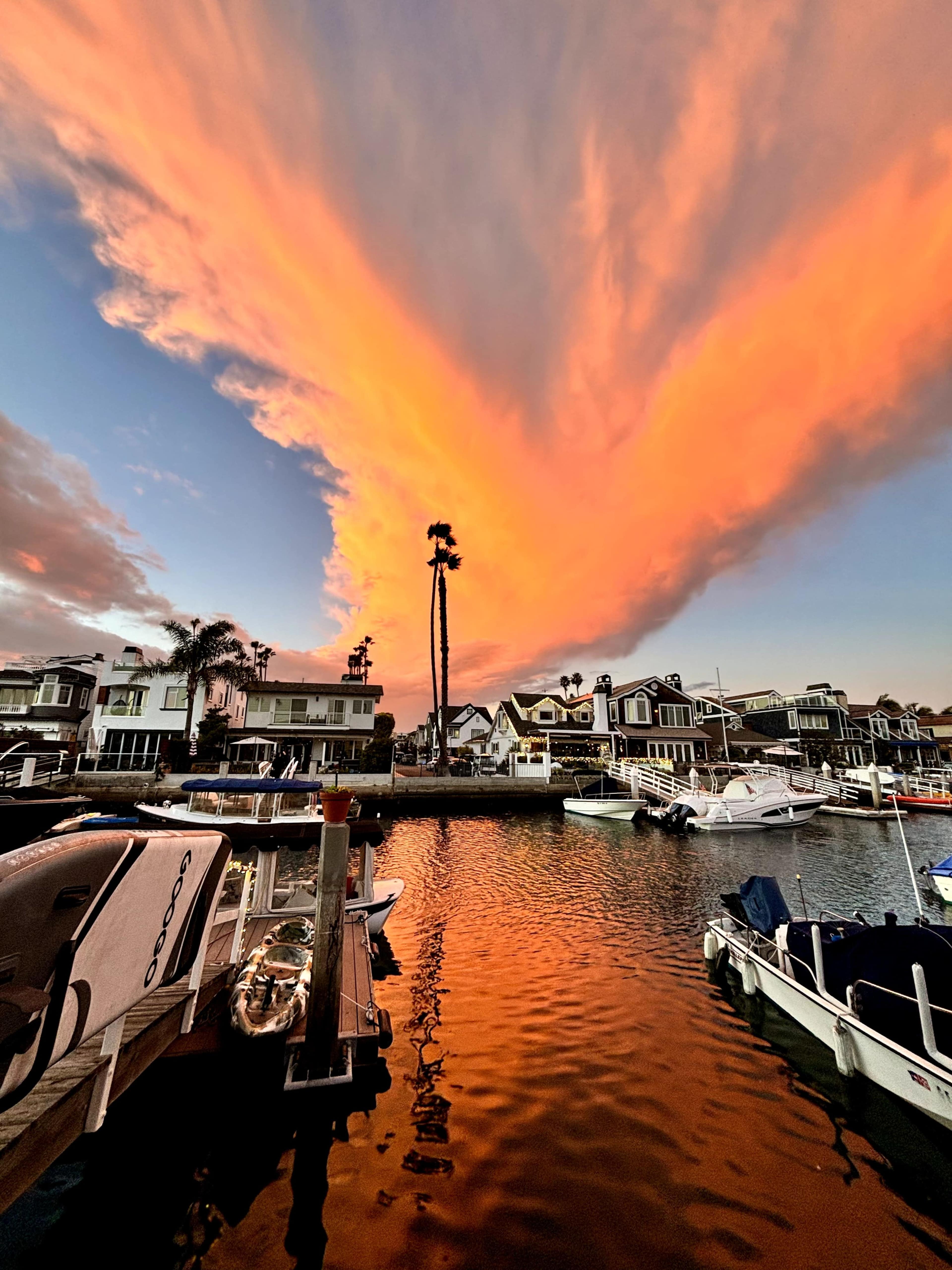 The image shows a waterfront scene with boats moored along a canal, flanked by houses, under a vibrant sky illuminated by orange and pink clouds.