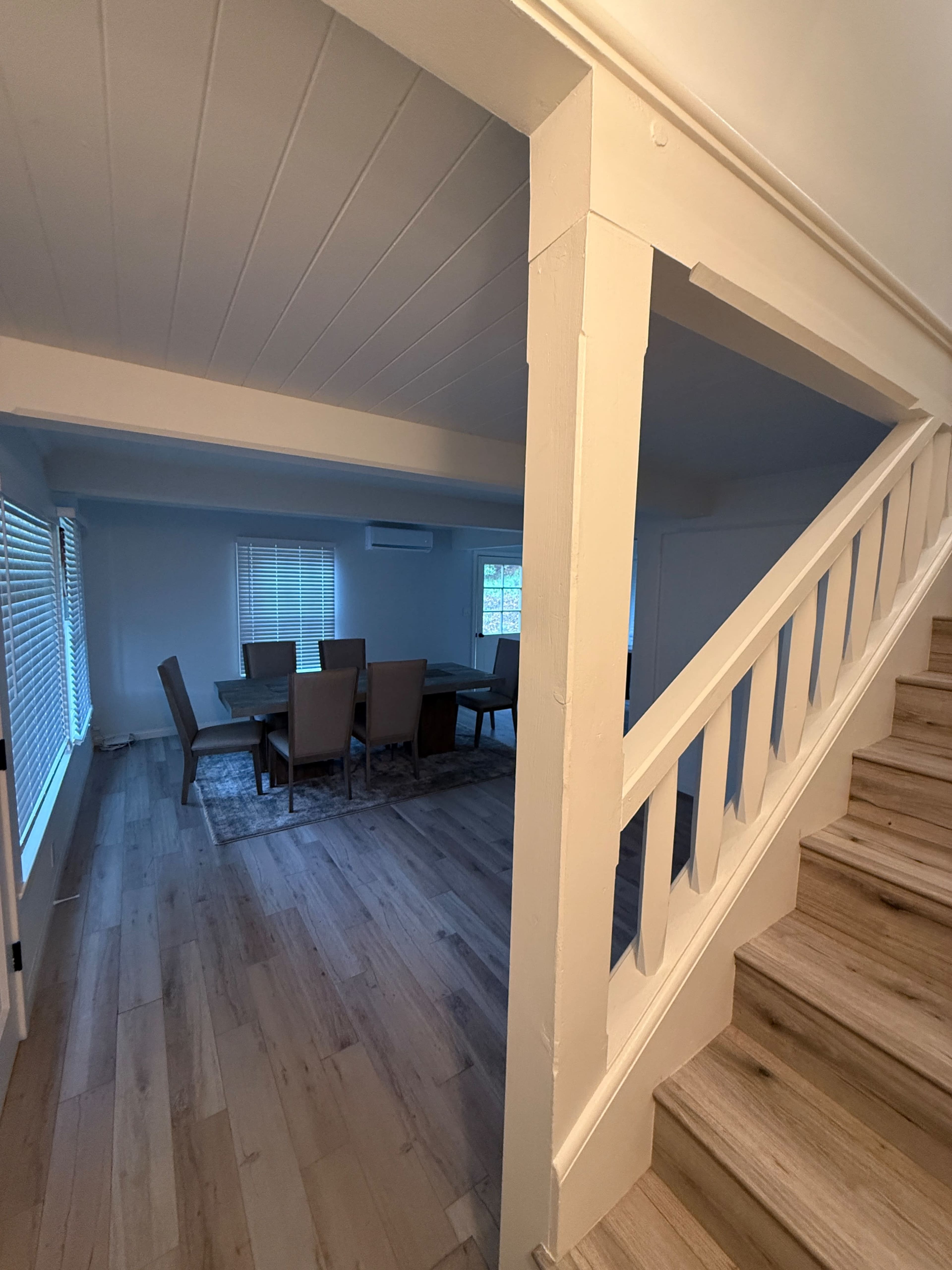 A staircase leads to a dining area with a large table and chairs, illuminated by natural light through windows.