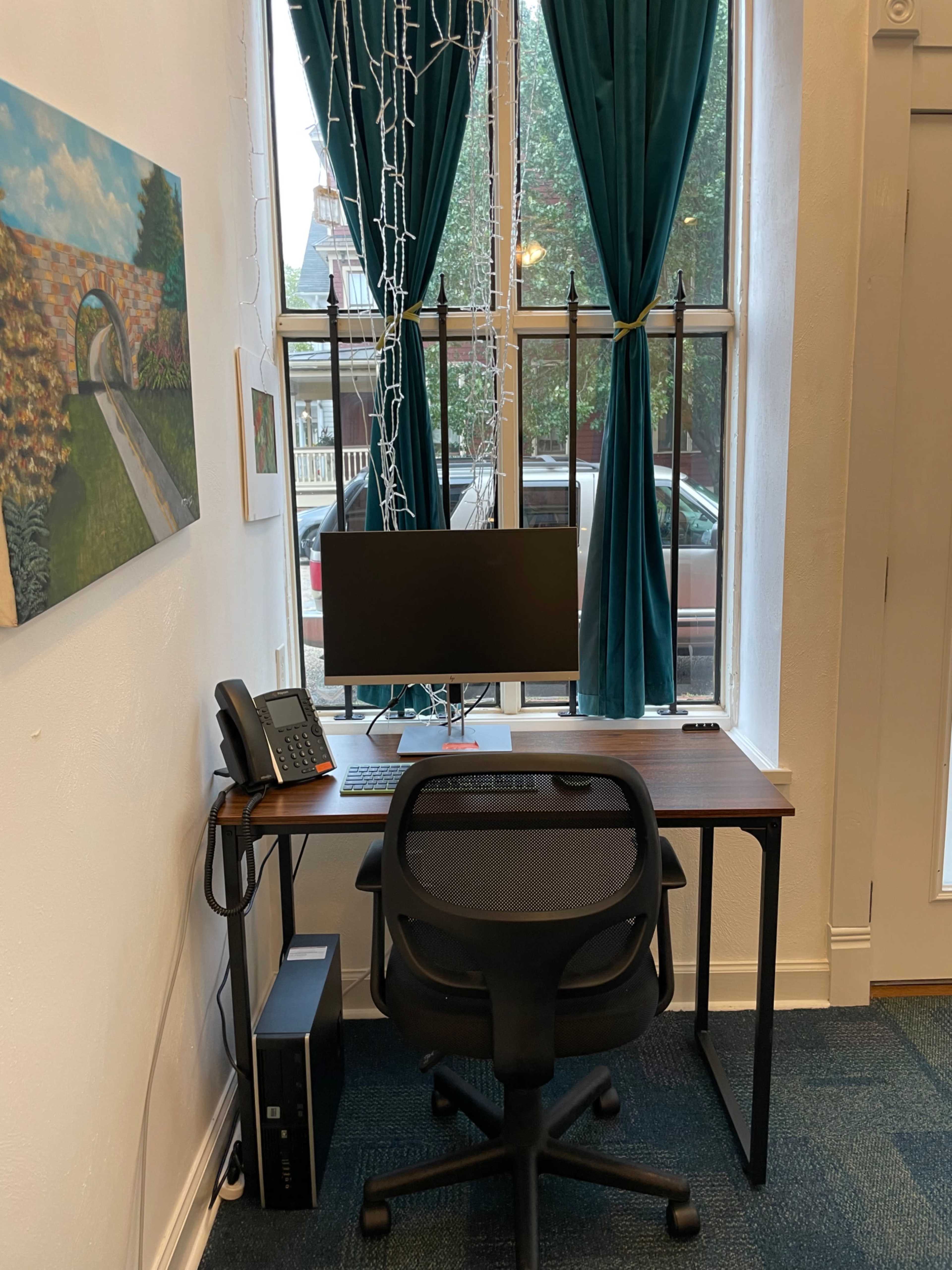 A modern desk with a computer monitor, telephone, and office chair sits next to a window draped with teal curtains.