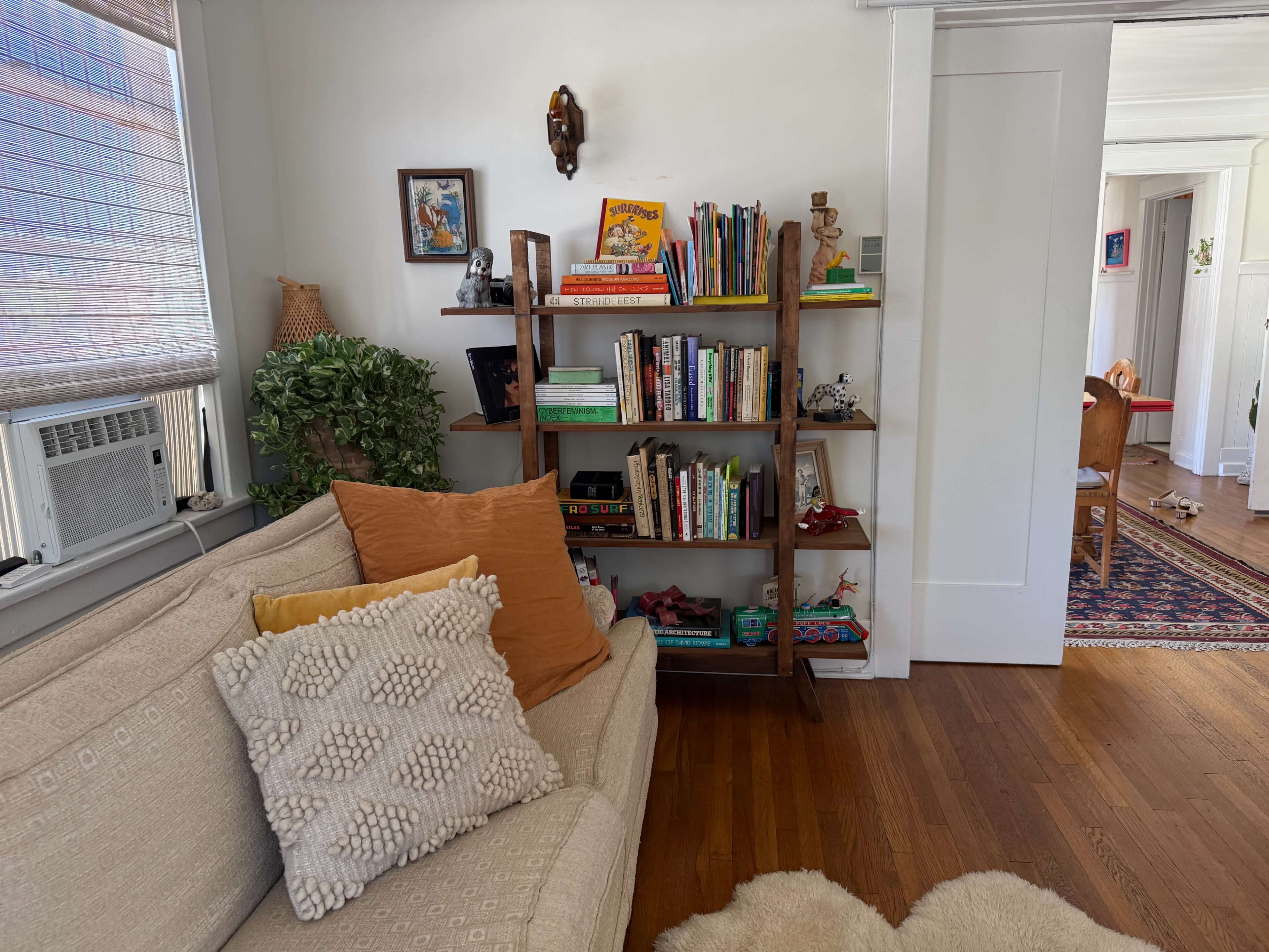 A wooden bookshelf filled with books and decorative items stands near a light-colored sofa with decorative pillows in a cozy room.