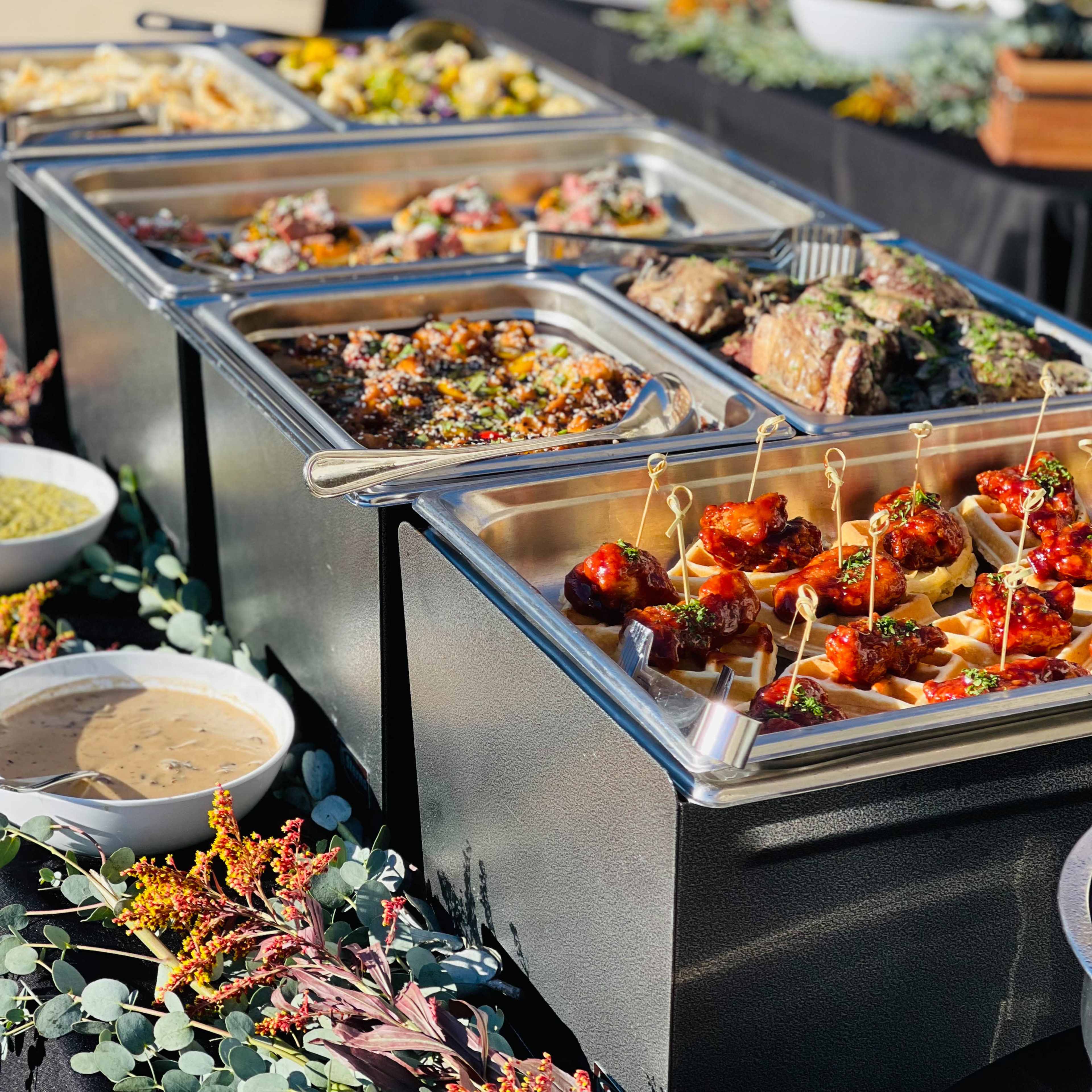 A buffet table displays various dishes, including meats, sauces, and appetizers, arranged in metal trays and surrounded by decorative foliage.