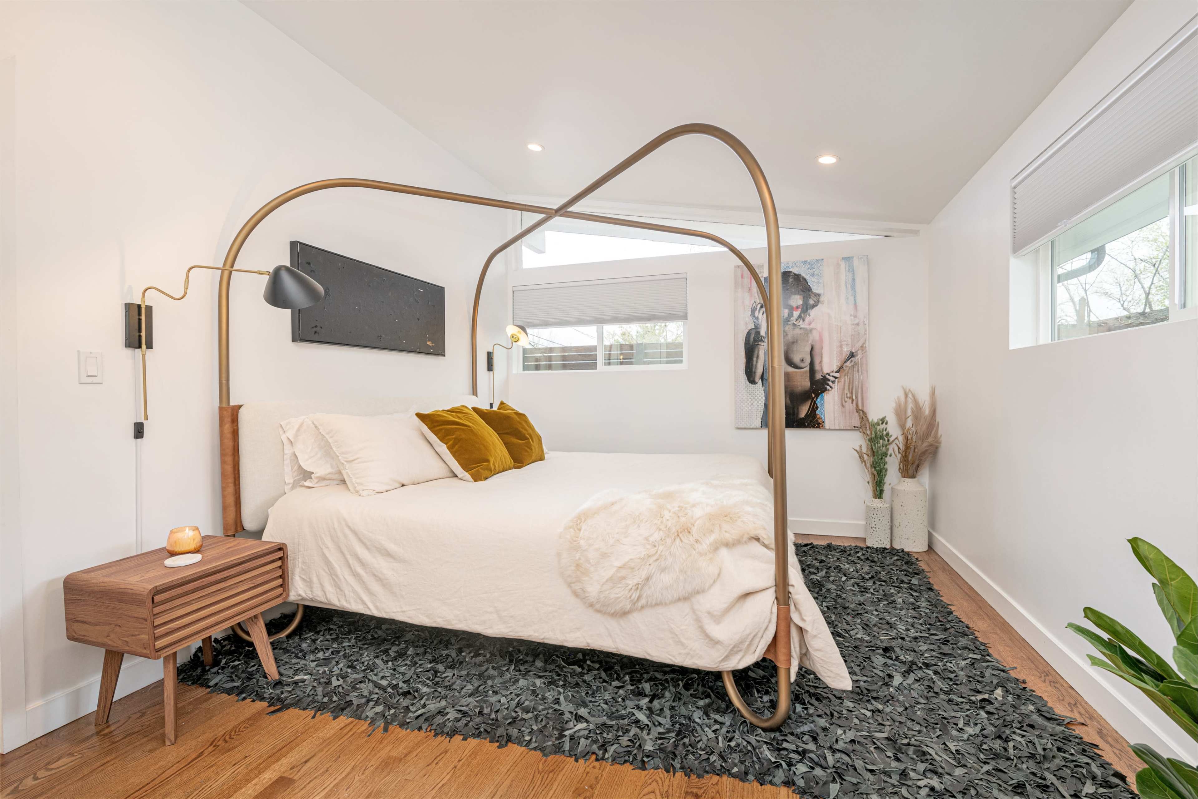 The bedroom features a modern bed with a unique metal frame, complemented by a wooden nightstand, a textured rug, and large windows.