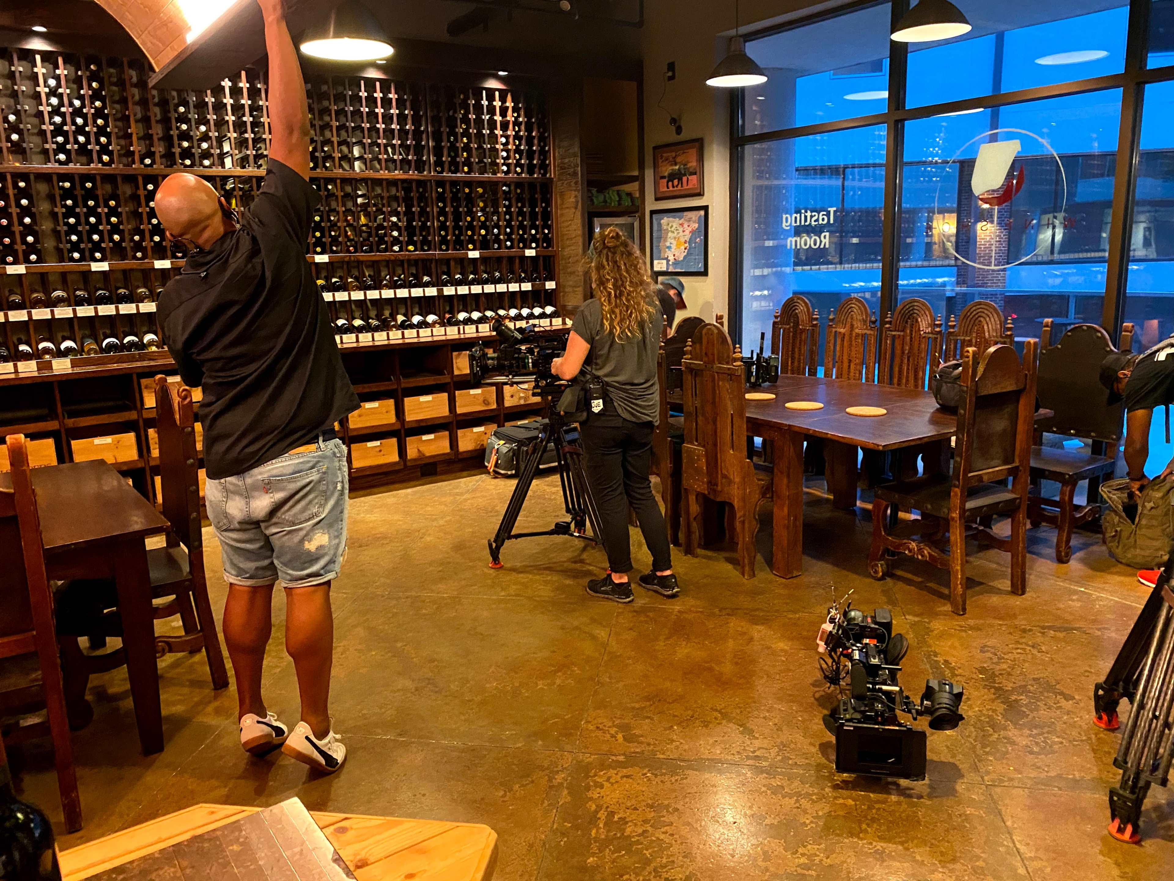 A man adjusts a light fixture while a woman sets up a camera in a restaurant with wooden furniture and a wall of wine bottles.