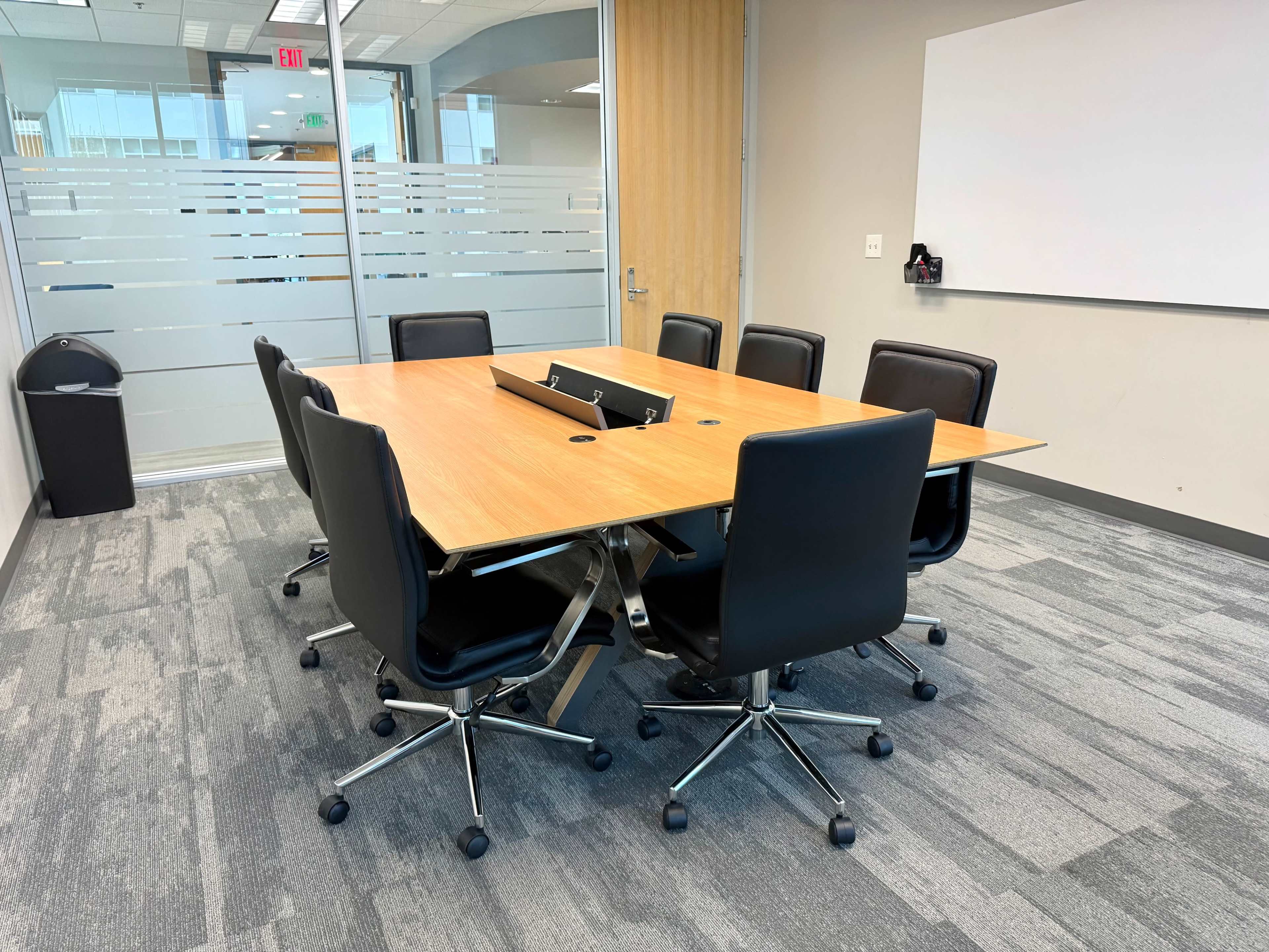 A conference room features a wooden table surrounded by six black ergonomic chairs, with glass partitions and a whiteboard visible in the background.