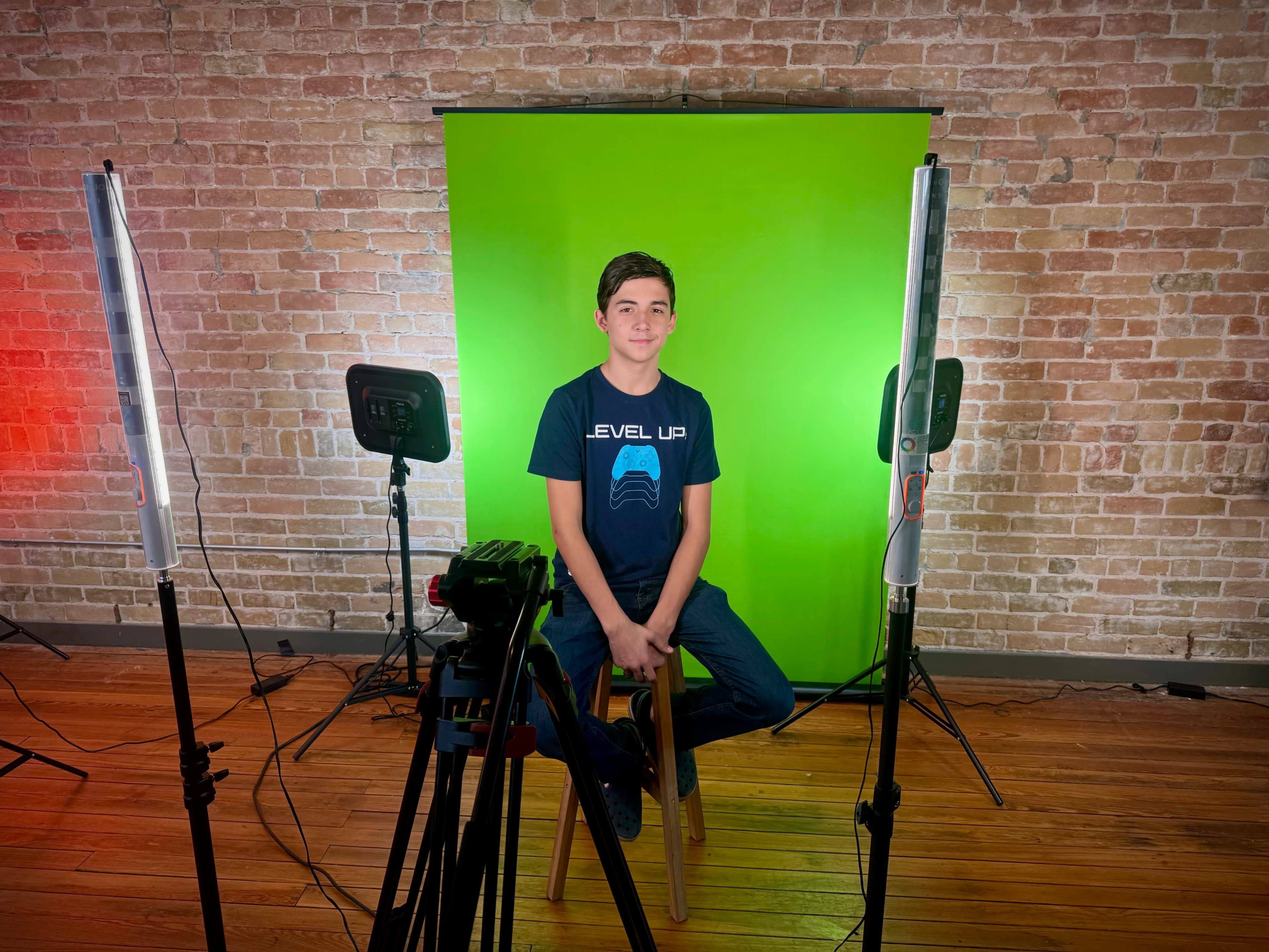 A boy sits on a stool in front of a green screen, surrounded by lighting equipment.