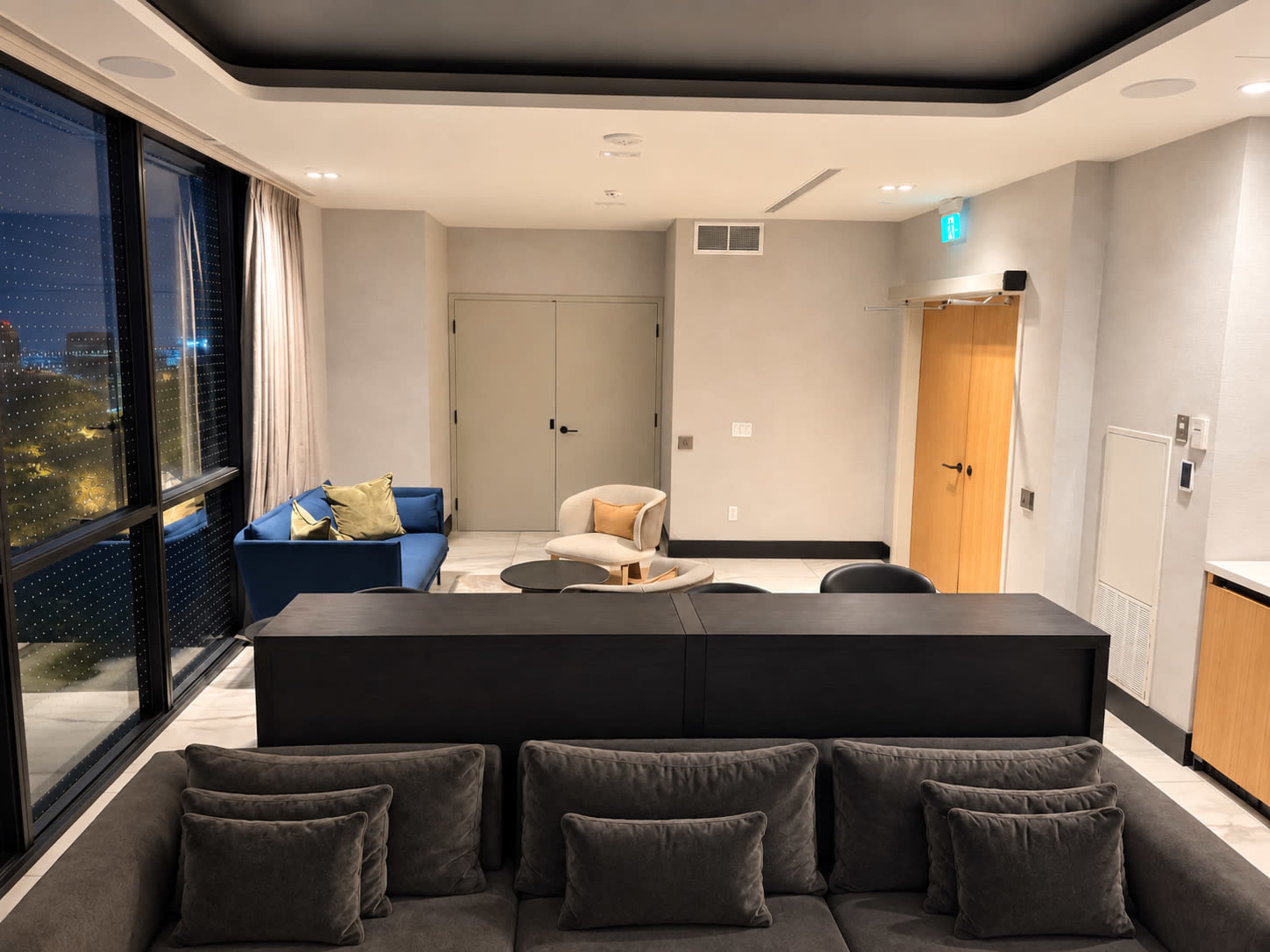 The image shows a modern lounge area with a dark sectional sofa, a round wooden coffee table, and a blue couch, all situated beside large windows displaying city views.