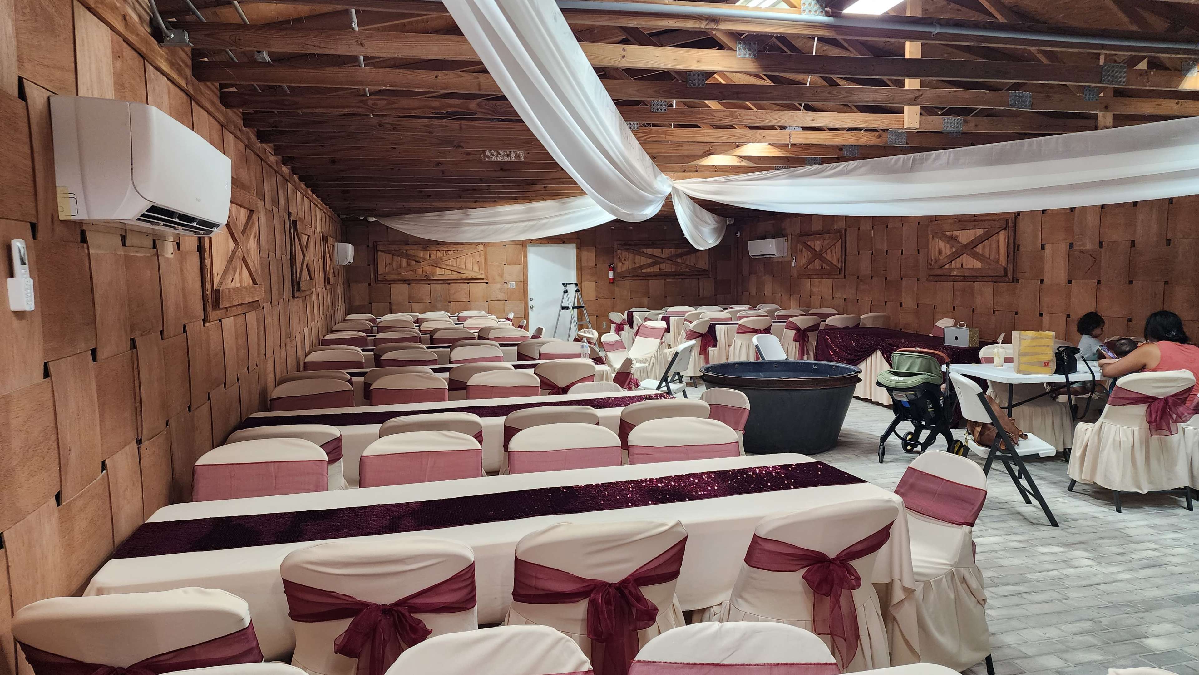 A decorated indoor event space with several rows of tables covered in white tablecloths and burgundy chair sashes, alongside wooden walls and overhead drapery.