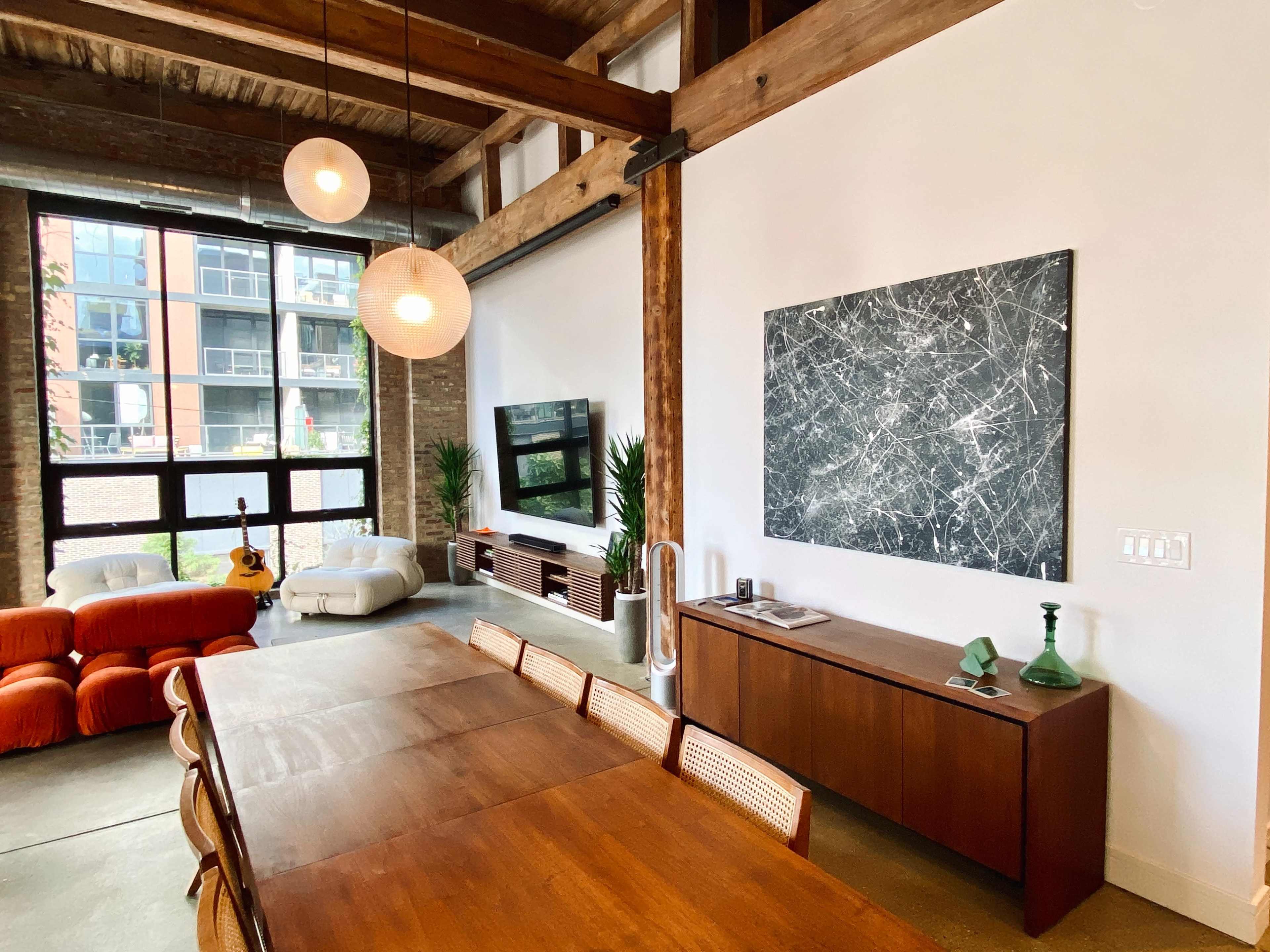 A modern dining area features a wooden table surrounded by chairs, with a large abstract painting on the wall and a view of a living space with a television and sofa.