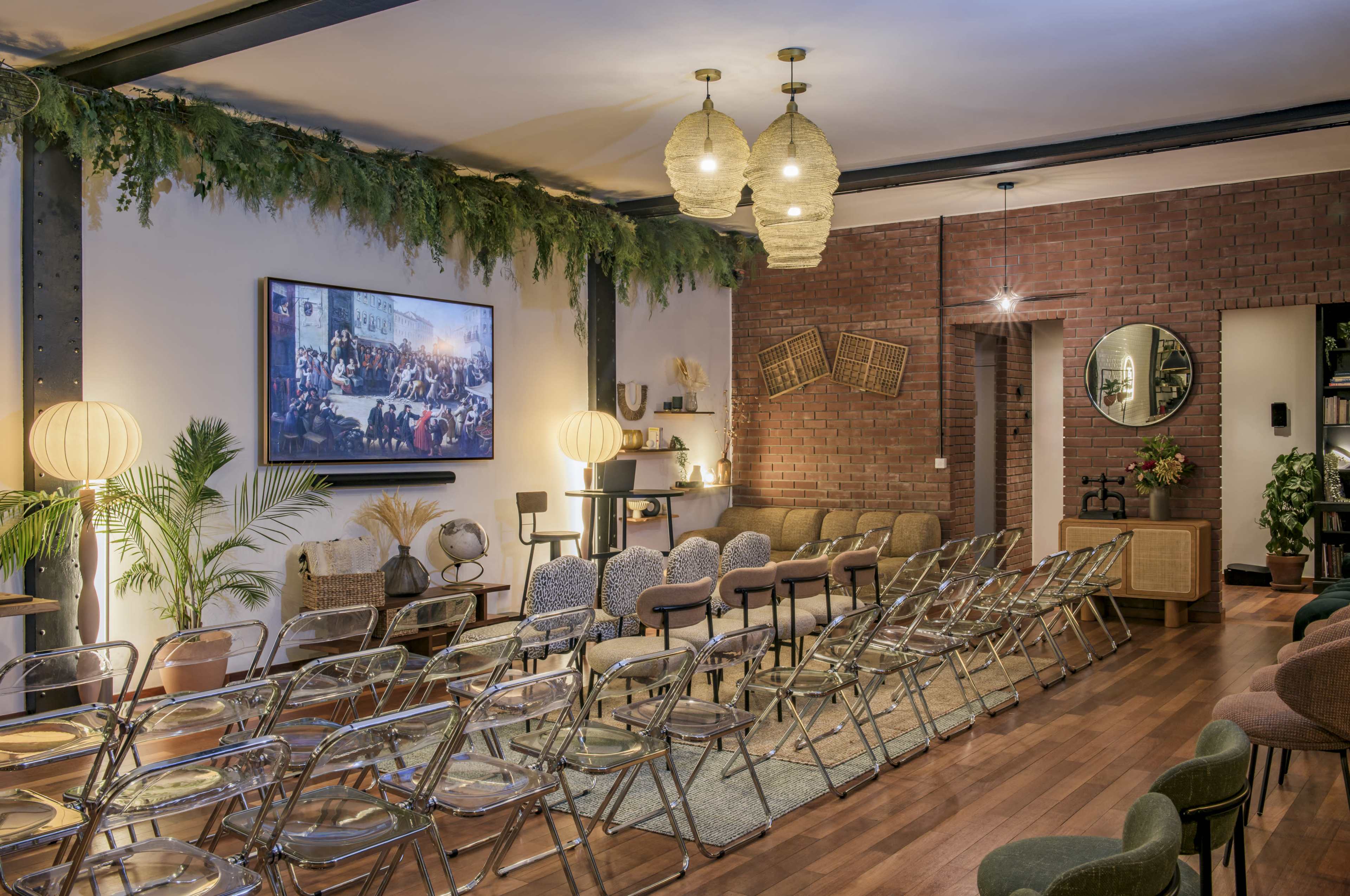 The image depicts a modern indoor event space with seating arranged in rows, decorative plants, soft lighting, and a large screen displaying an image on the wall.