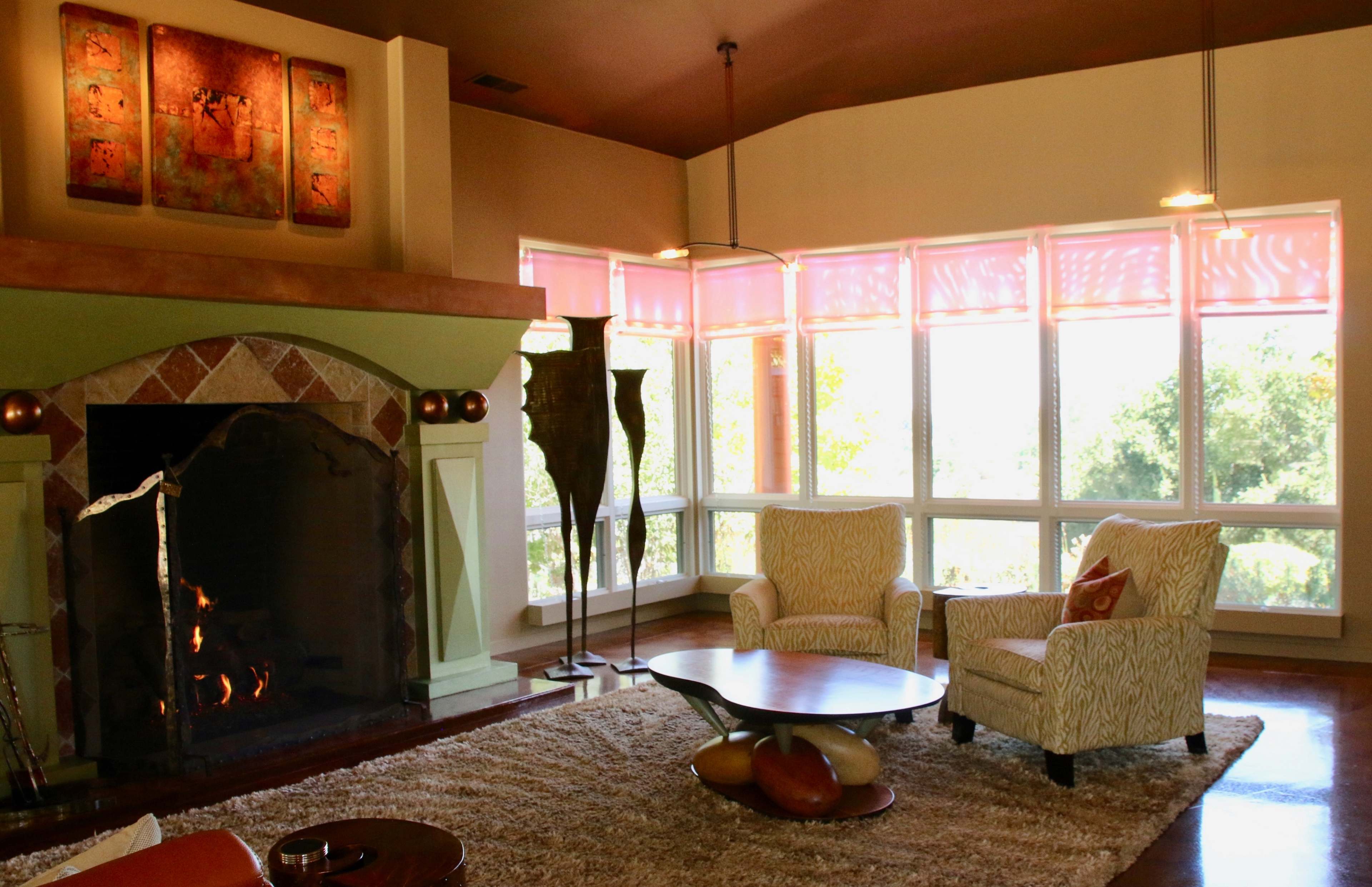 The scene features a cozy living room with a large fireplace, two armchairs, a circular coffee table, and large windows allowing natural light.