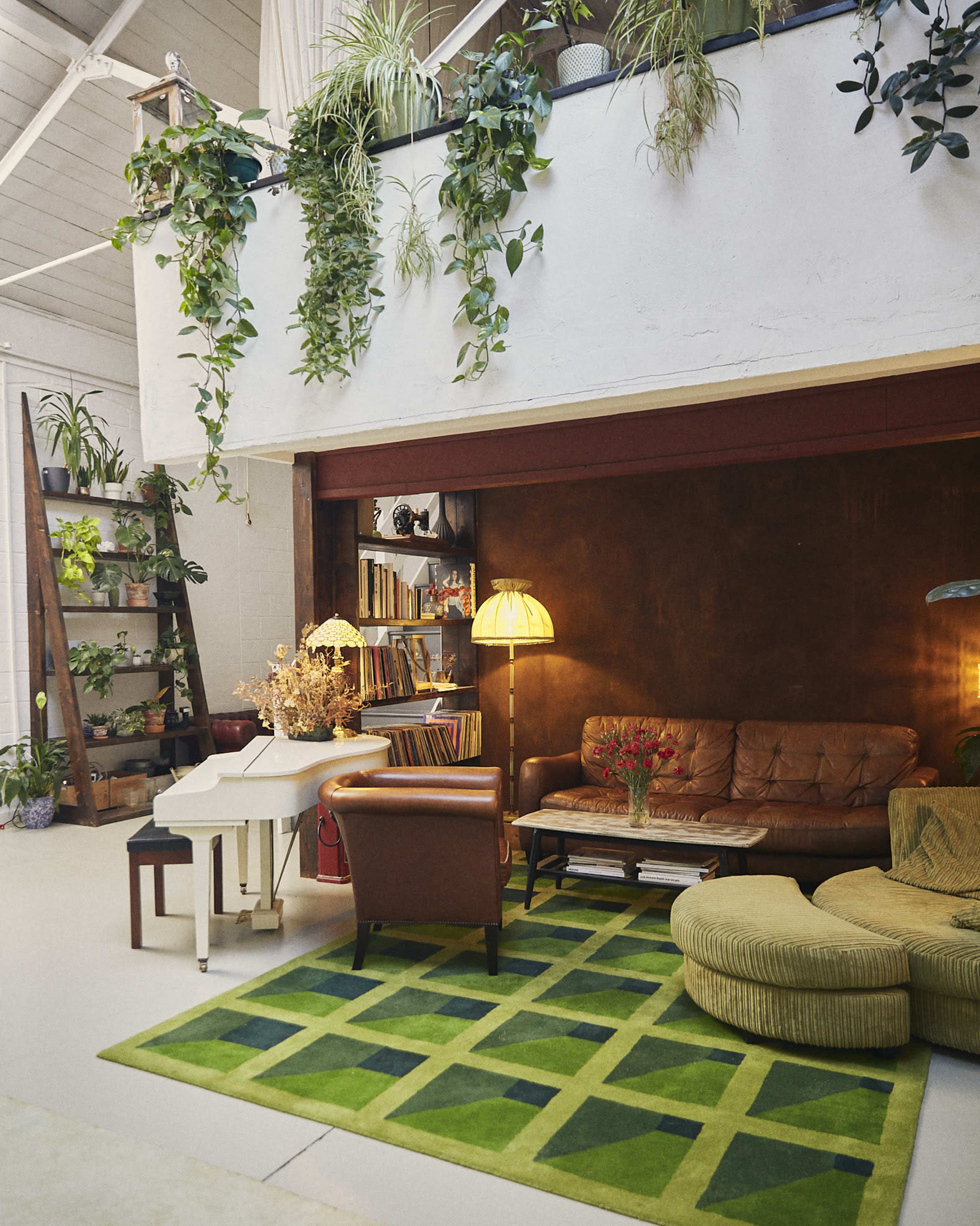 The image depicts a modern living space with a brown leather sofa, a green patterned rug, a warm lamp, and potted plants lining the shelves and walls.