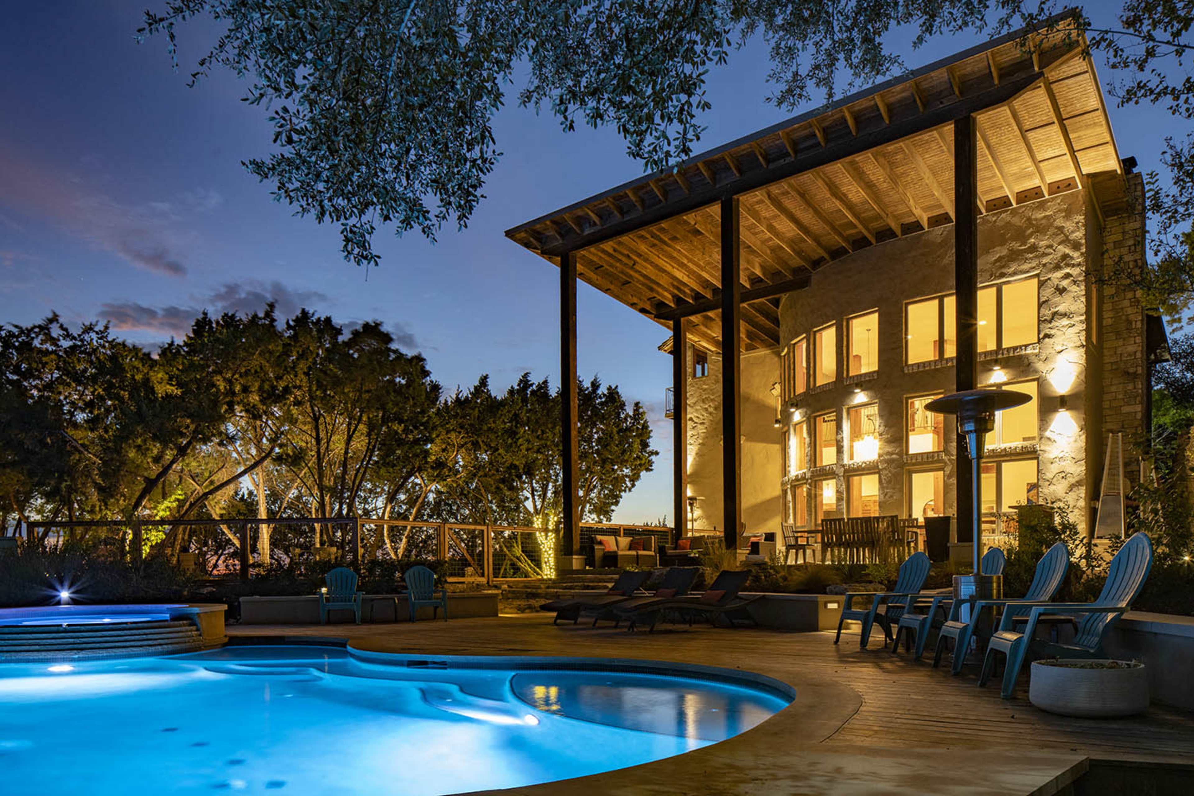 The image shows a modern two-story house with large windows, situated beside a pool and surrounded by trees in a twilight setting.