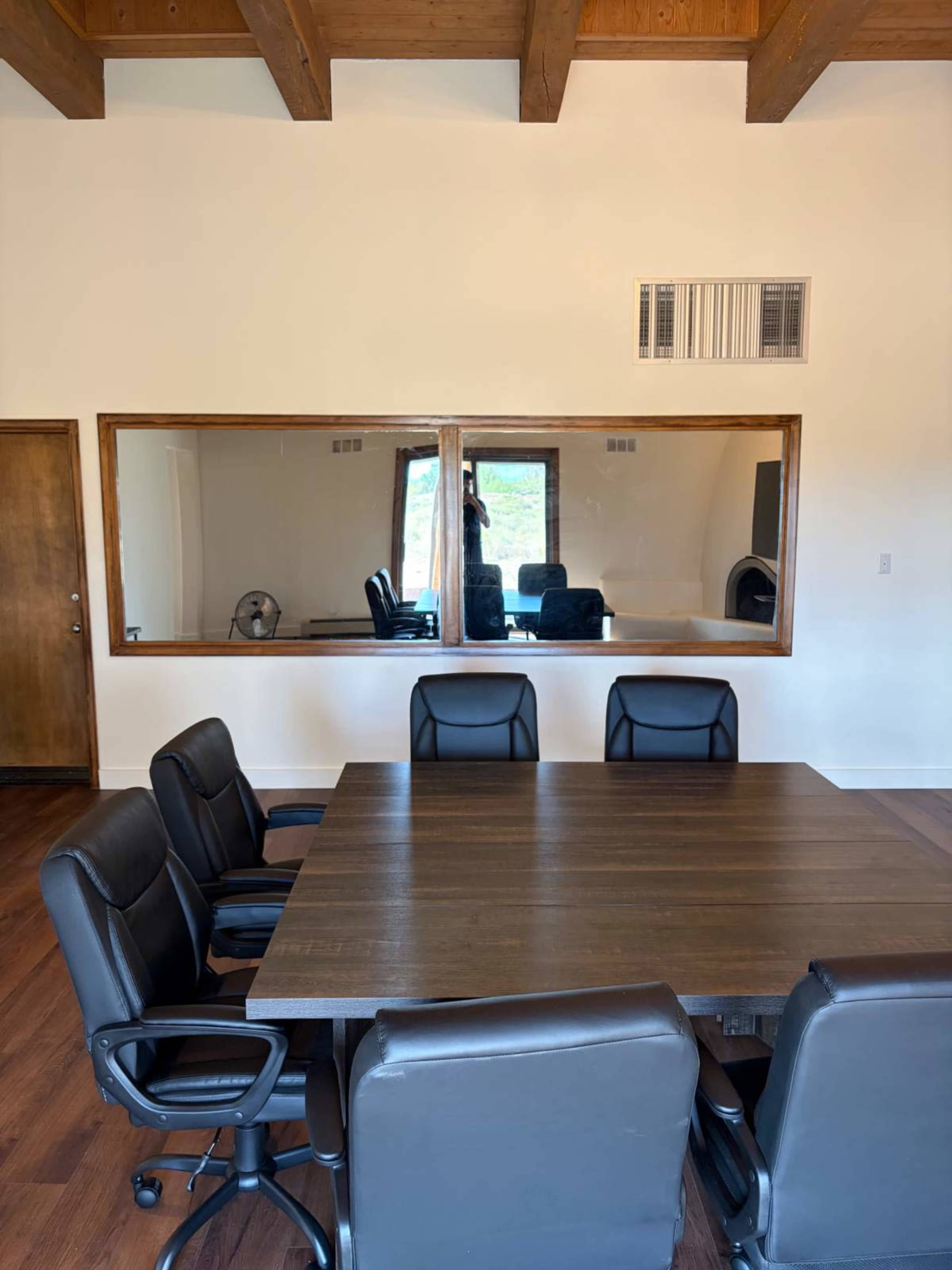 A conference room features a large wooden table surrounded by black ergonomic chairs, with a mirrored wall showcasing a view of another space.