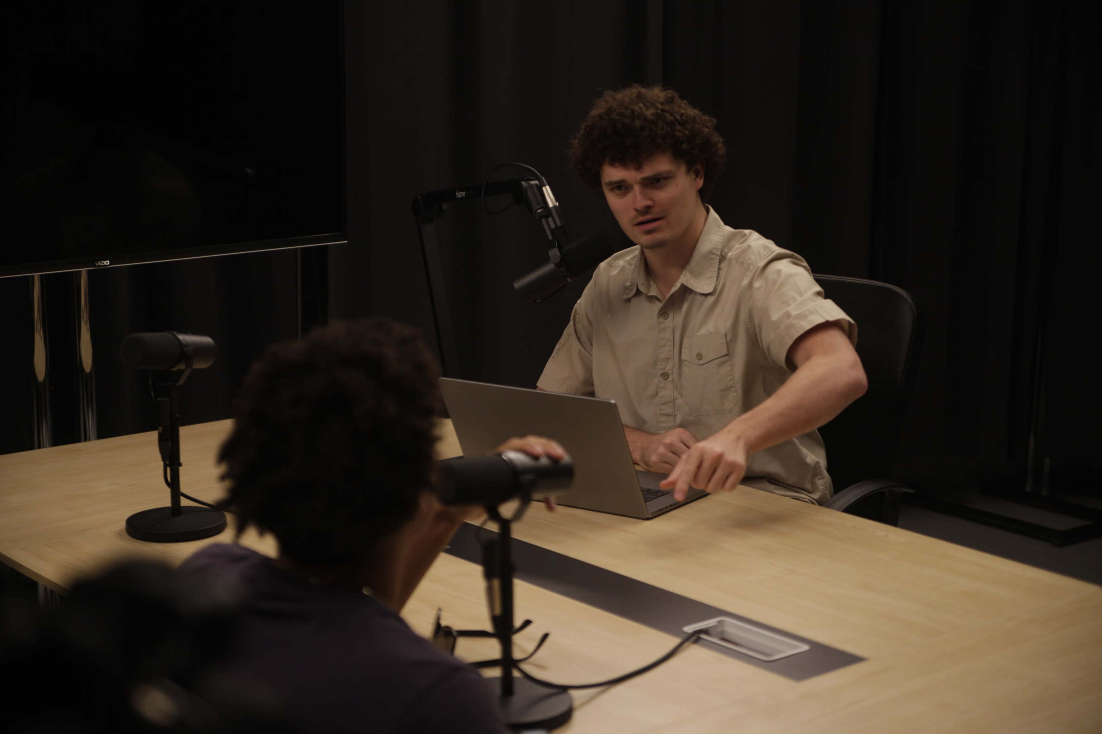 Two individuals are seated at a table with a laptop and microphones, engaged in a discussion in a dimly lit studio.