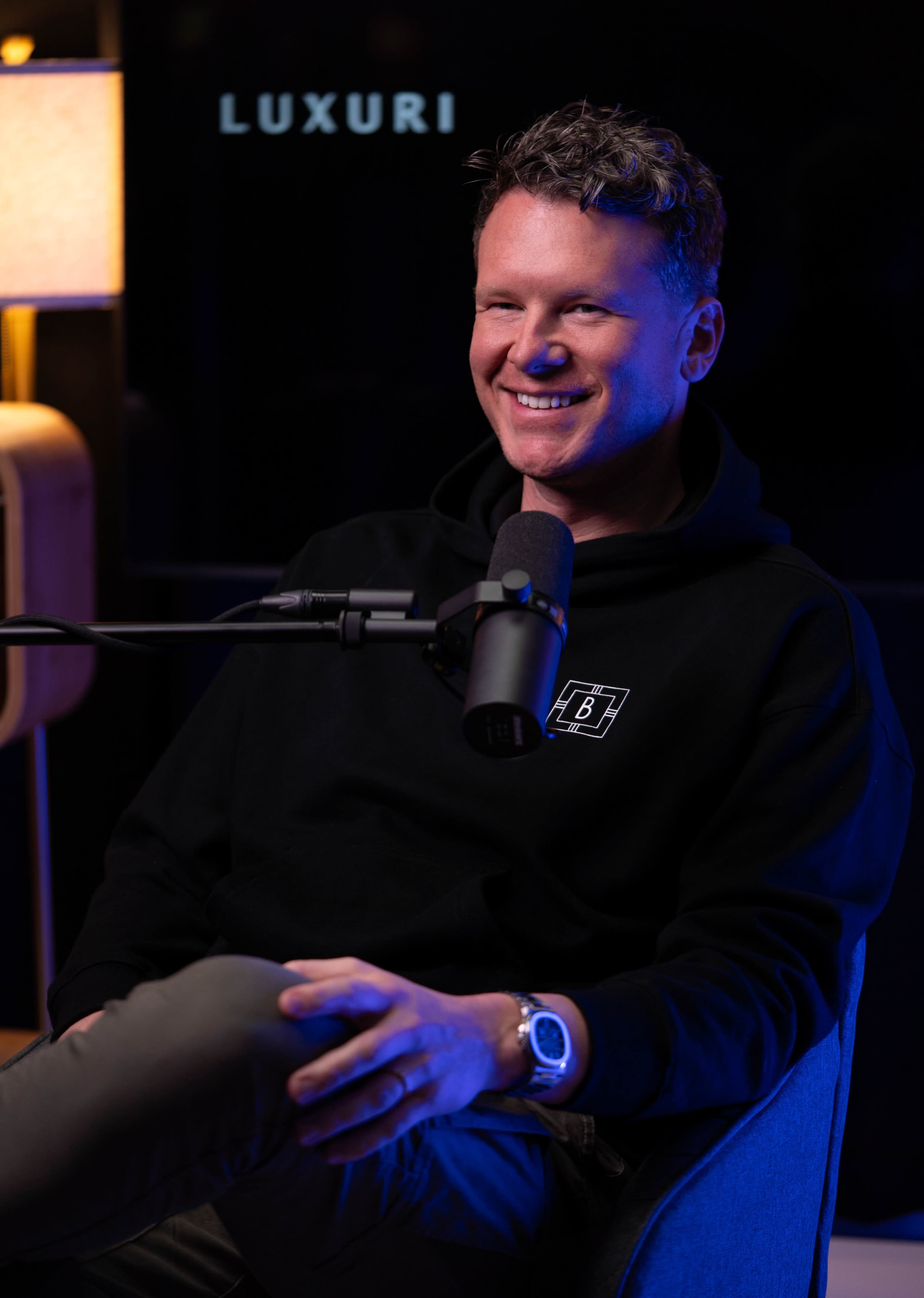 A man with curly hair is sitting in a chair, wearing a black hoodie and smiling while holding a microphone in a modern studio setting.