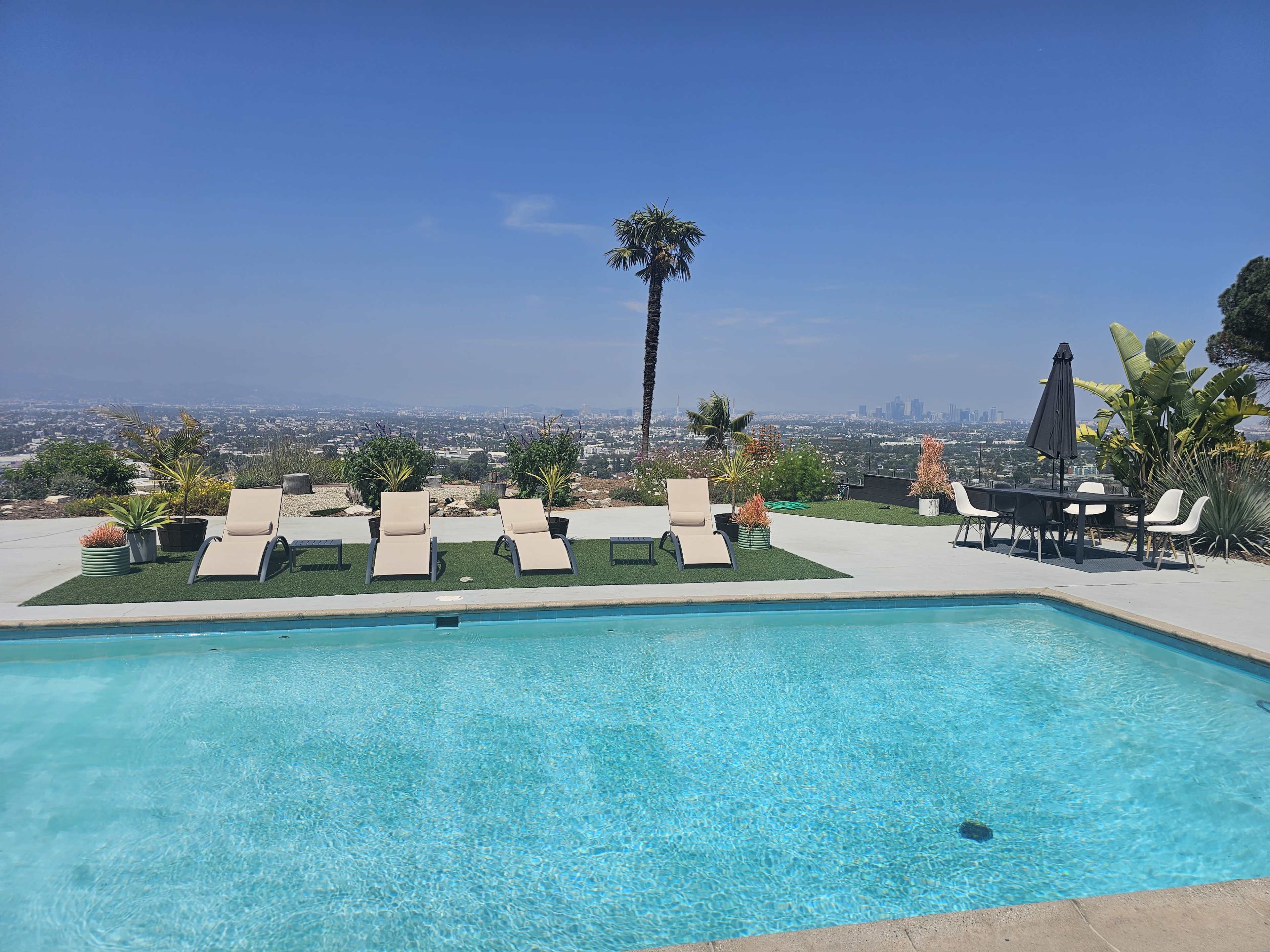 A clear swimming pool with lounge chairs and a dining table overlooks a city skyline surrounded by greenery.