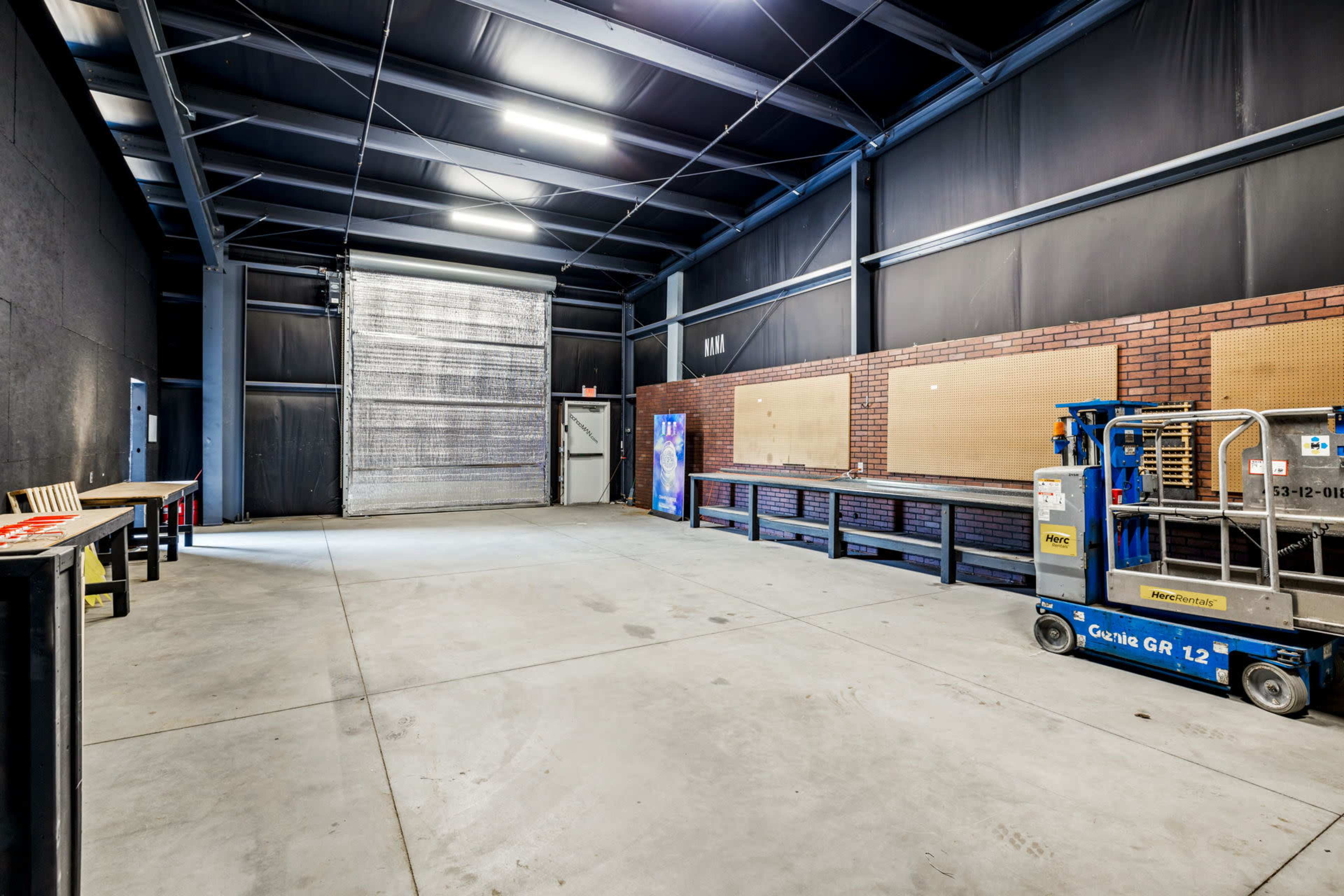 An empty industrial space with high ceilings, concrete flooring, and large rolling doors, along with work tables and a lift in one corner.
