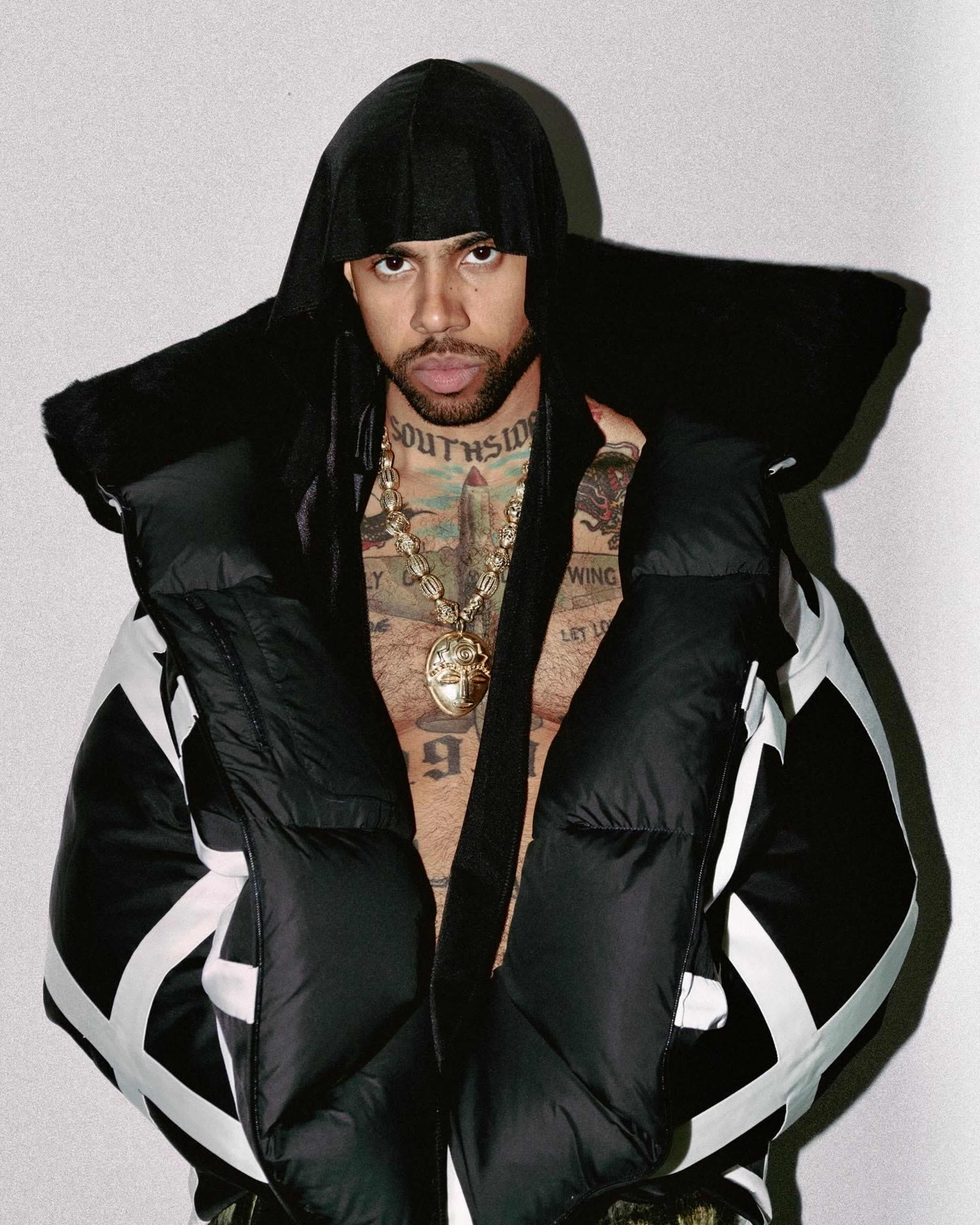 A man with tattoos is wearing a large black puffer jacket and a head covering, standing against a plain background.
