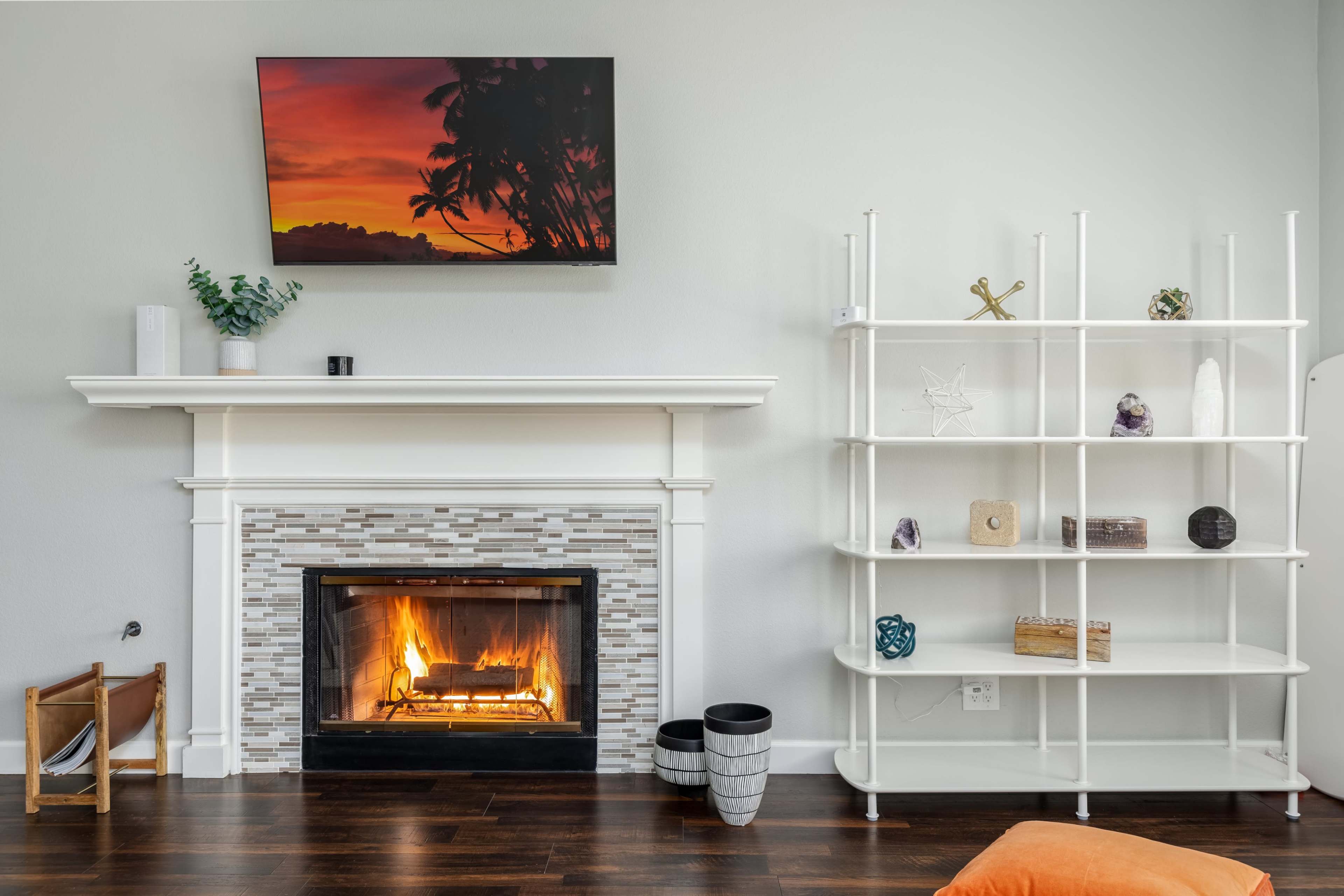 A modern living room features a fireplace with a mosaic surround, a white shelving unit, and a television mounted on the wall displaying a sunset.