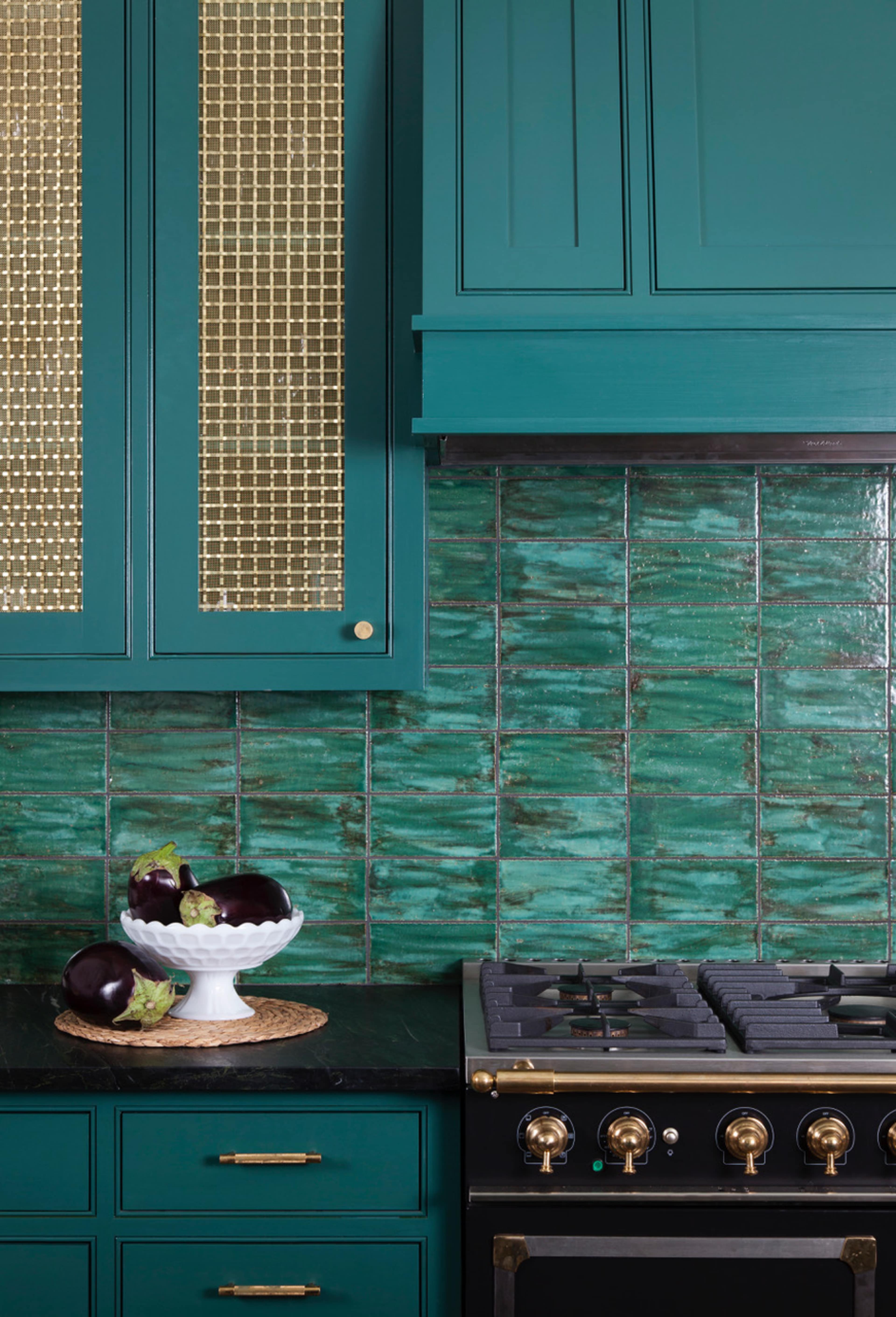 The image shows a kitchen with teal cabinetry, gold accents, and patterned tile backsplash, featuring a gas stove and a bowl of eggplants on the counter.