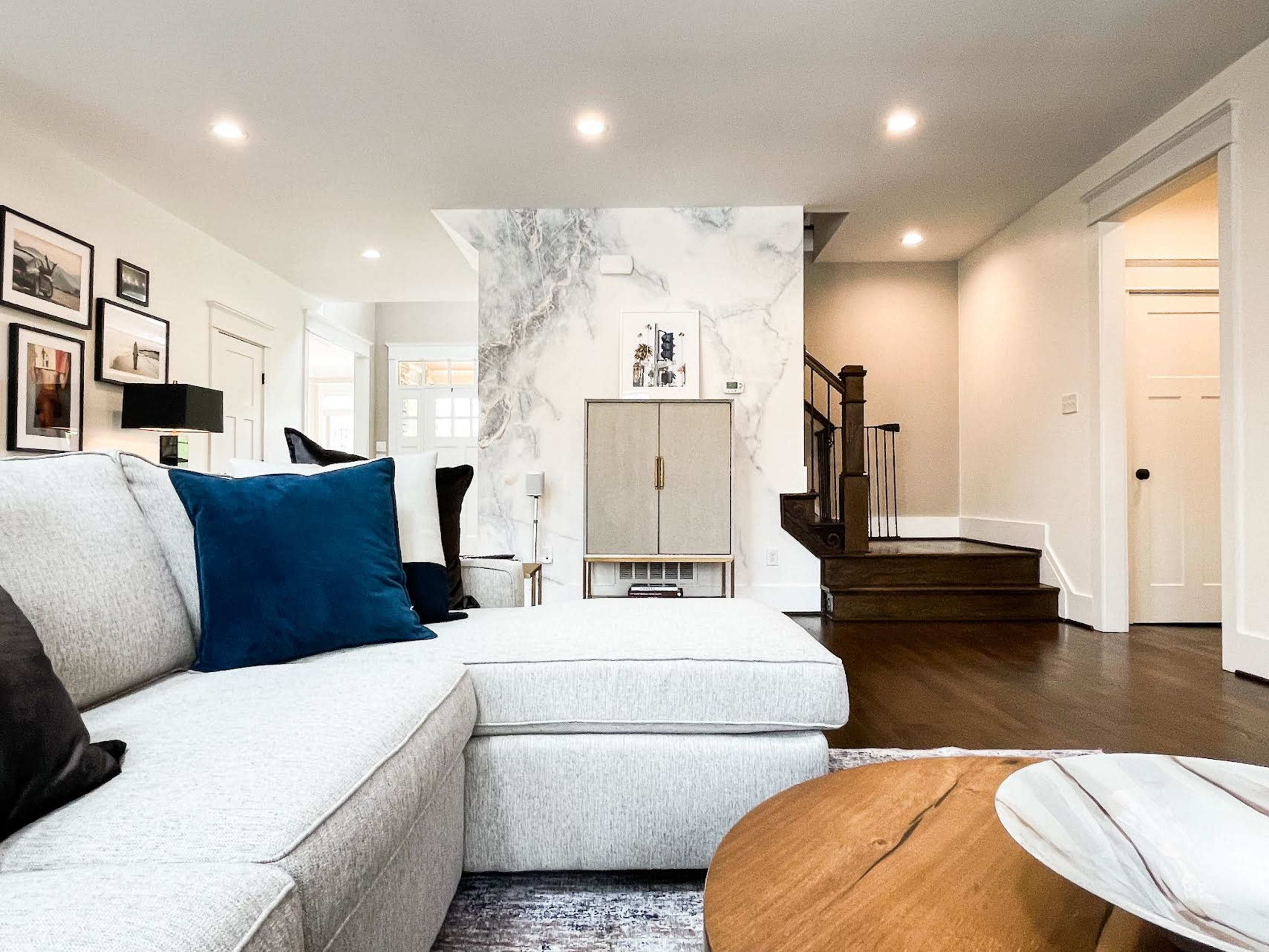 A modern living room with a light-colored sectional sofa, a wooden coffee table, and a staircase leading to another level, accented by a marble wall.