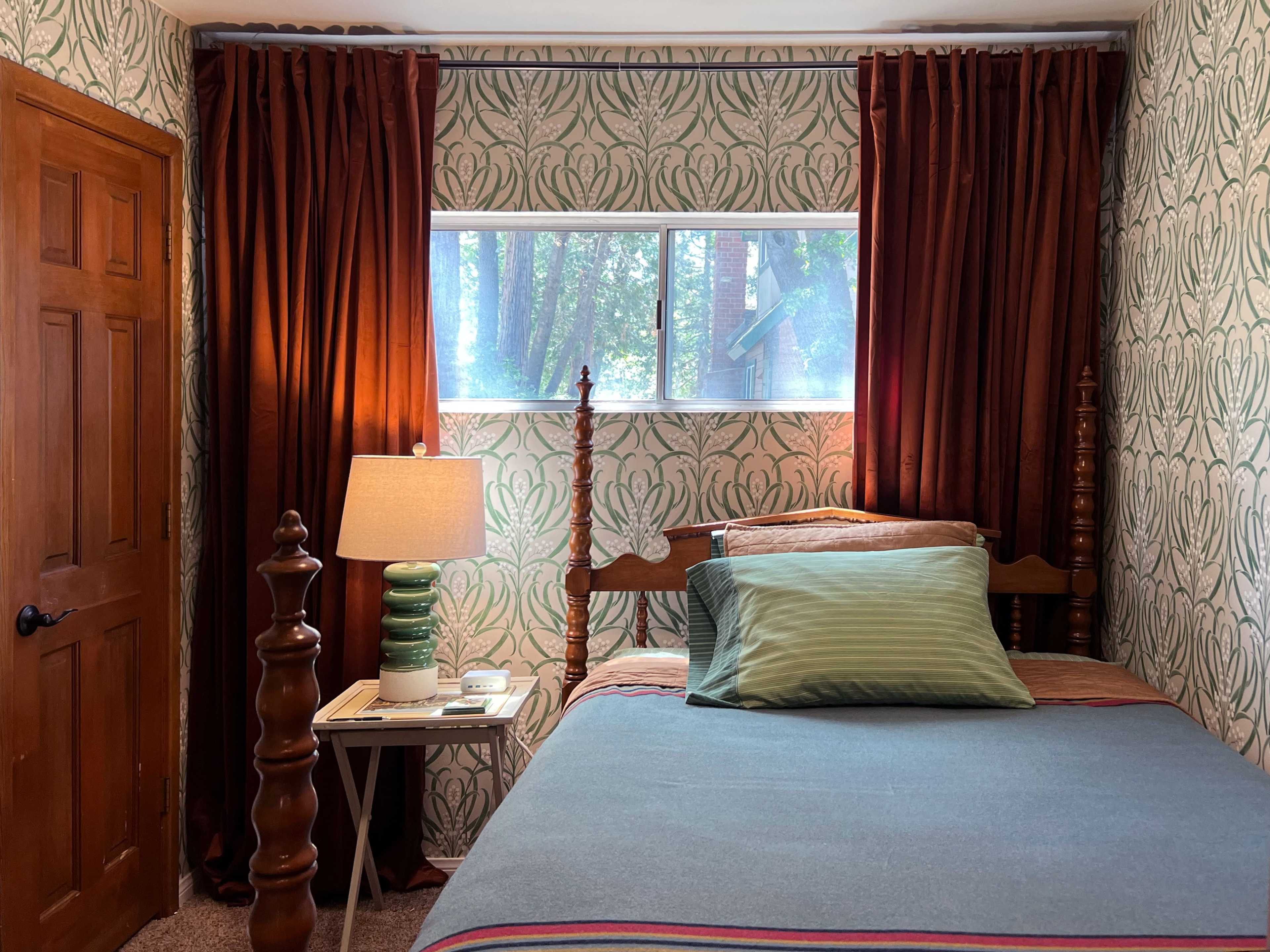 A small bedroom features a single bed with a green and striped blanket, a bedside lamp on a table, and patterned wallpaper with a window showing greenery outside.
