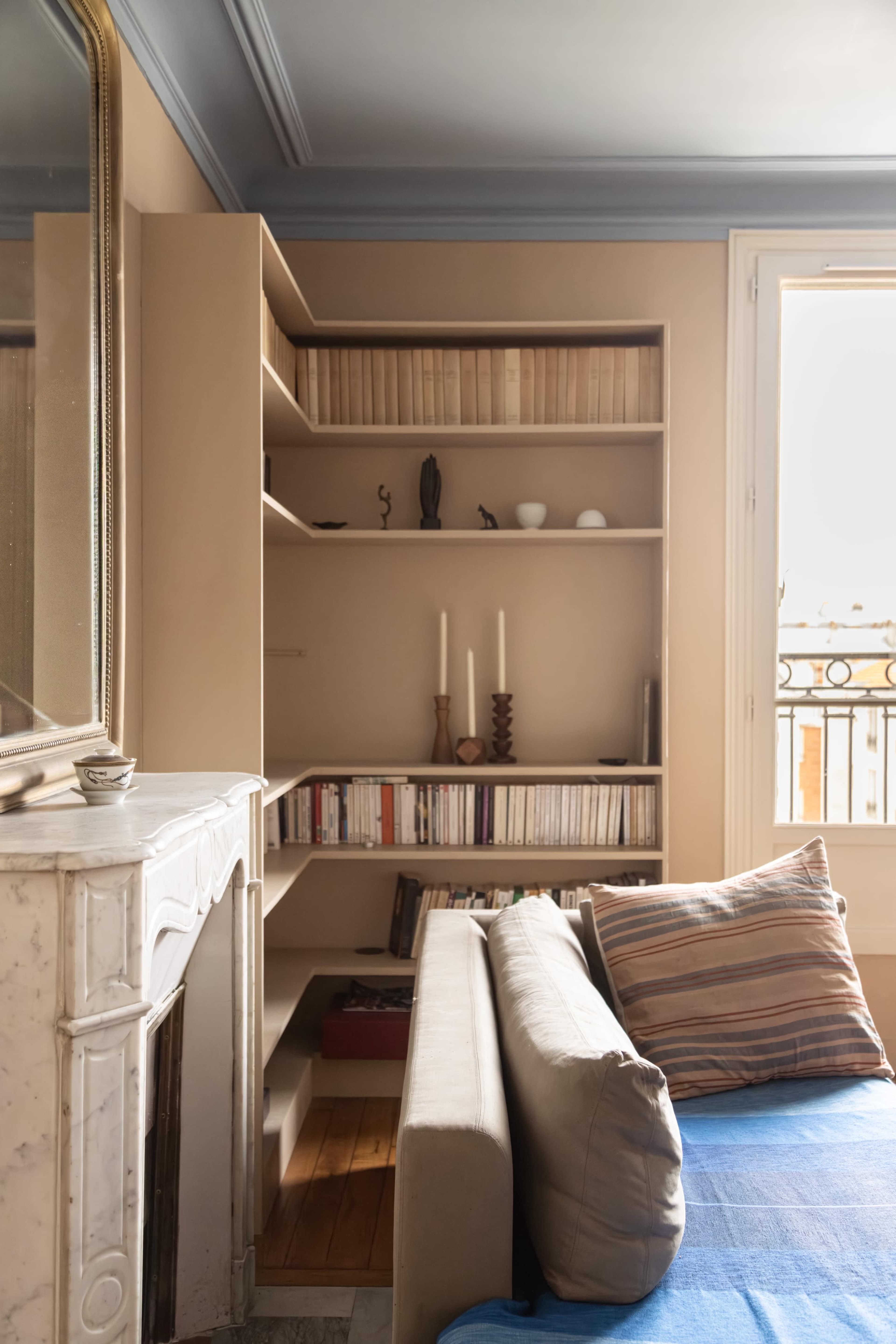 A cozy room features a modern bookshelf filled with books and decorative items, a gray sofa, and a marble fireplace beneath a large mirror, with natural light streaming in through a window.