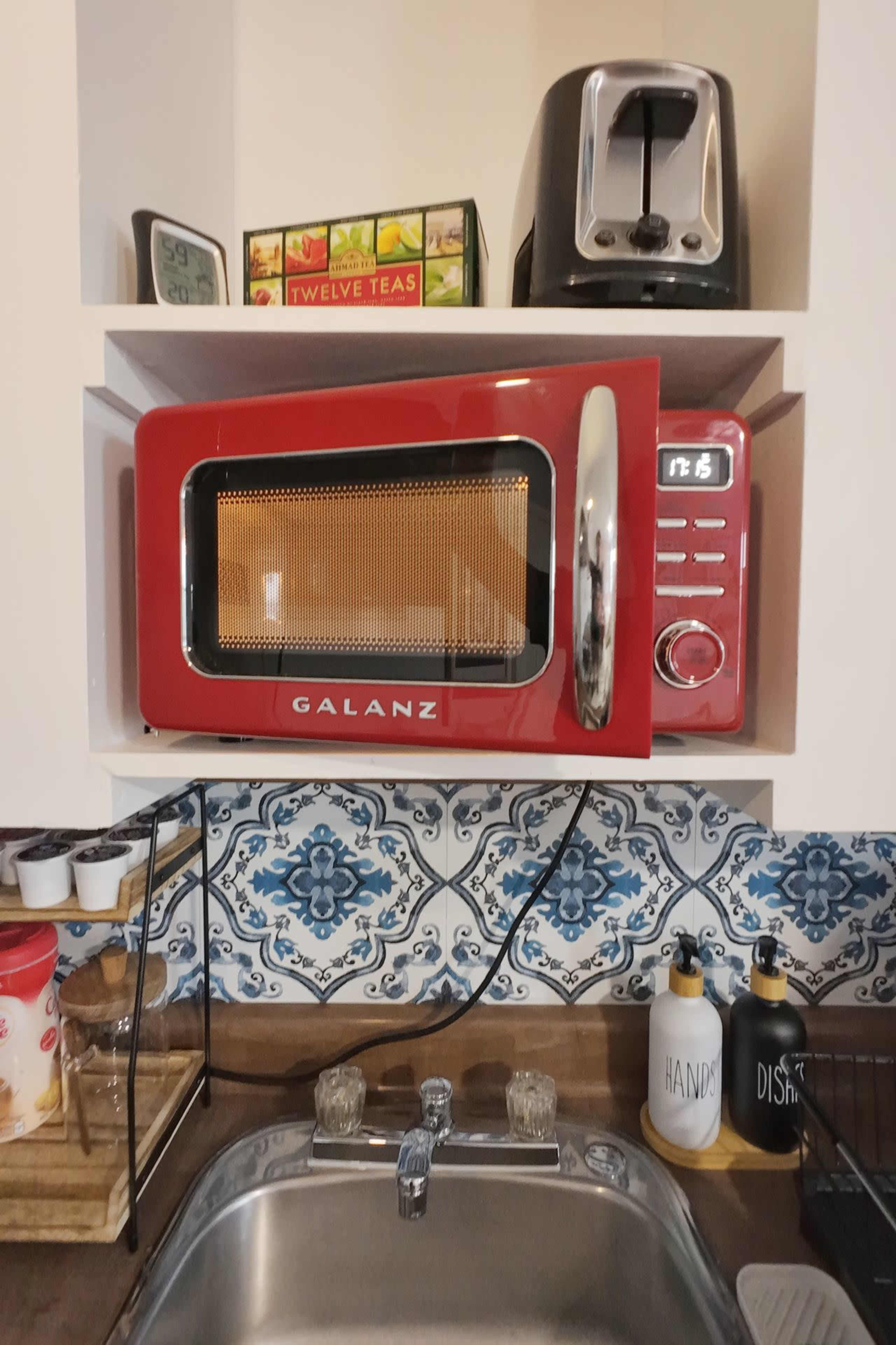 The image shows a red Galanz microwave placed on a shelf above a kitchen sink, with a toaster and tea boxes on the shelf above it.