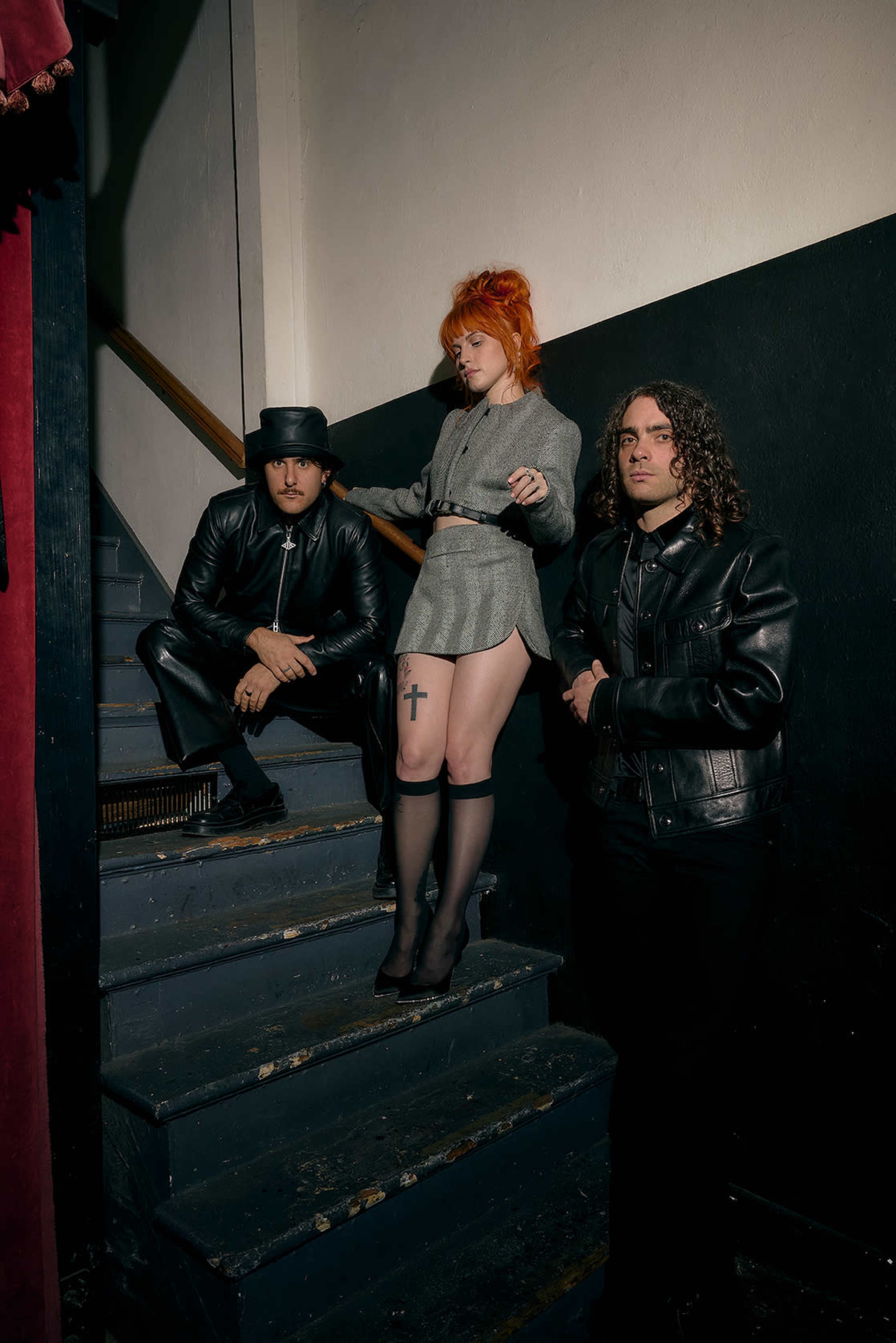 Three individuals pose on a staircase, with one sitting on the steps and two standing, dressed in dark fashion styles.