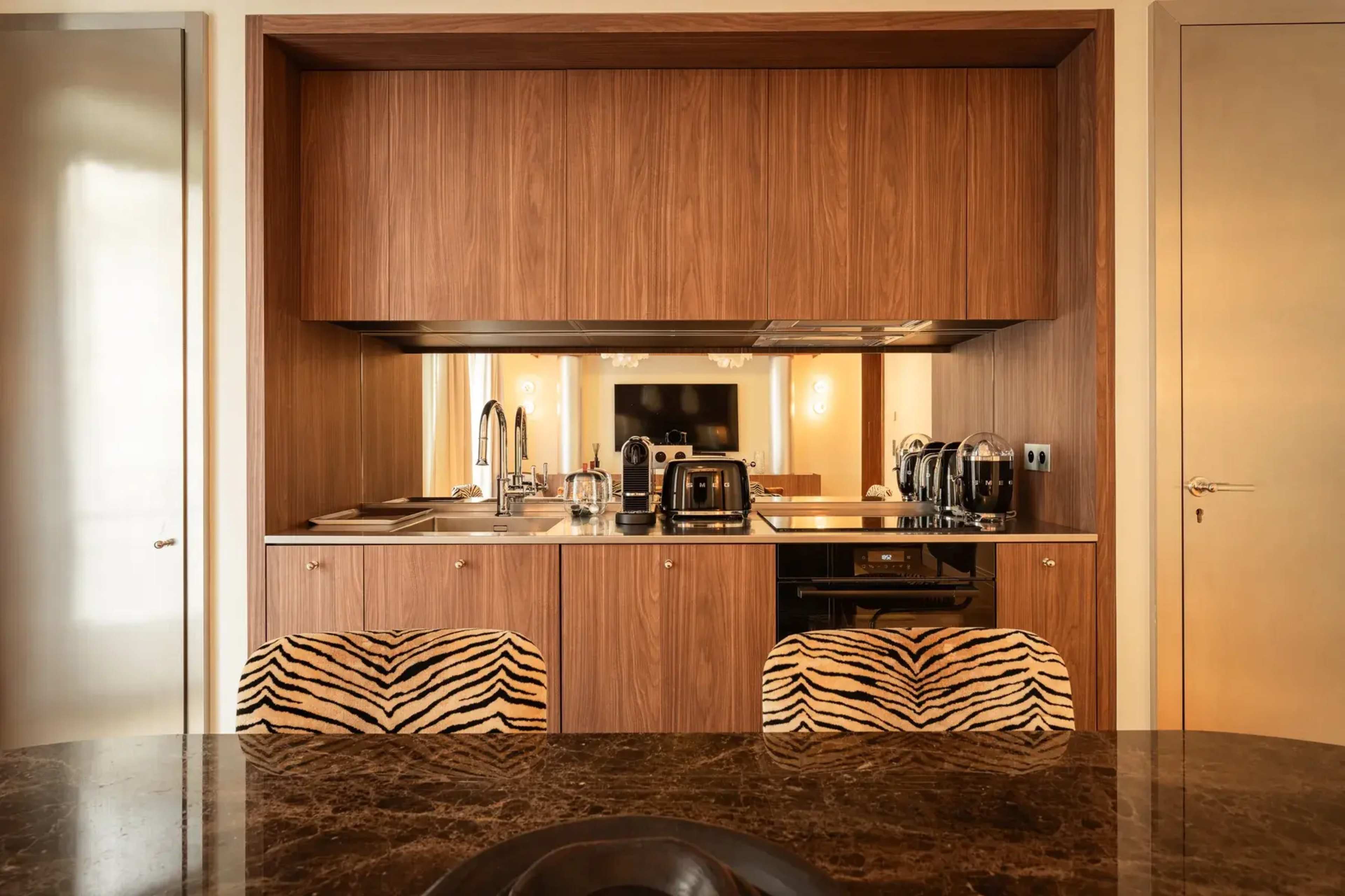 The image shows a modern kitchen area with a wooden cabinetry backdrop, featuring stainless steel appliances, and two chairs with a zebra print pattern at a marble dining table.