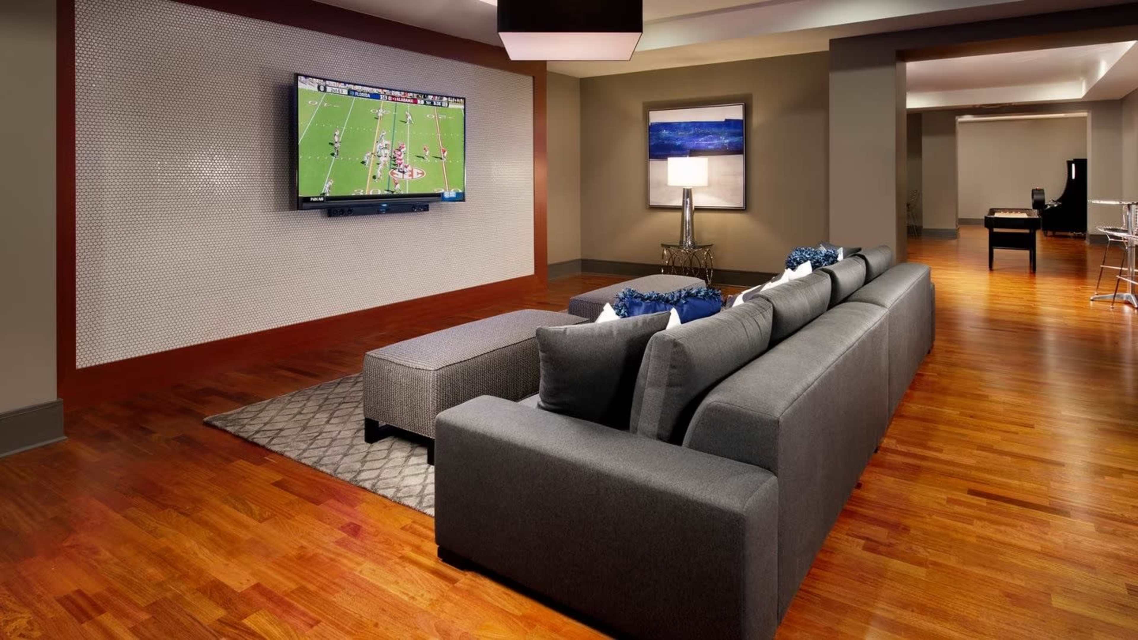 A modern living room features a gray sectional sofa facing a wall-mounted television displaying a football game, with hardwood flooring and minimalist decor.