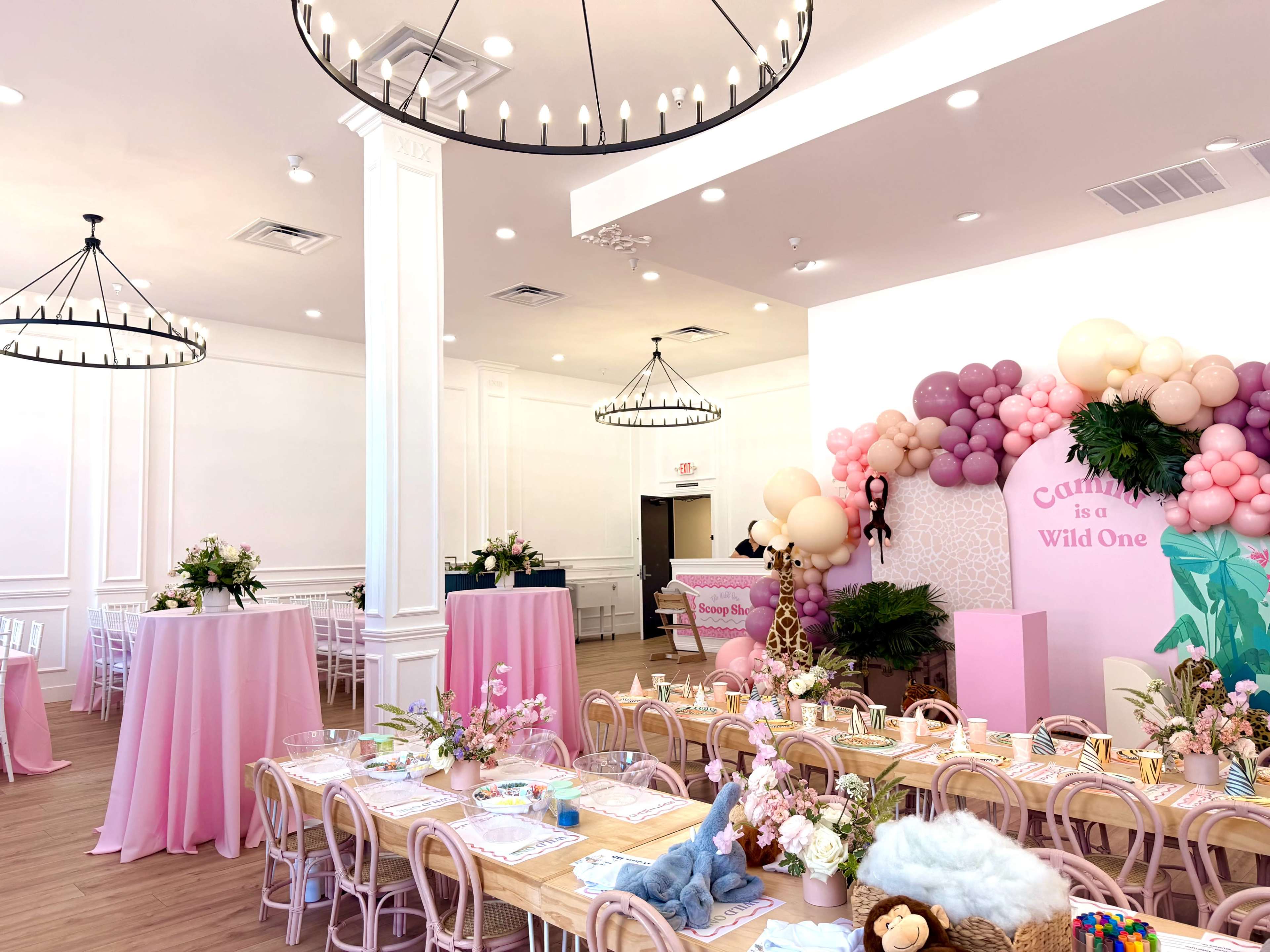 The image shows a decorated event space featuring tables set with floral arrangements and pastel-colored balloons, with a whimsical theme for a celebration.