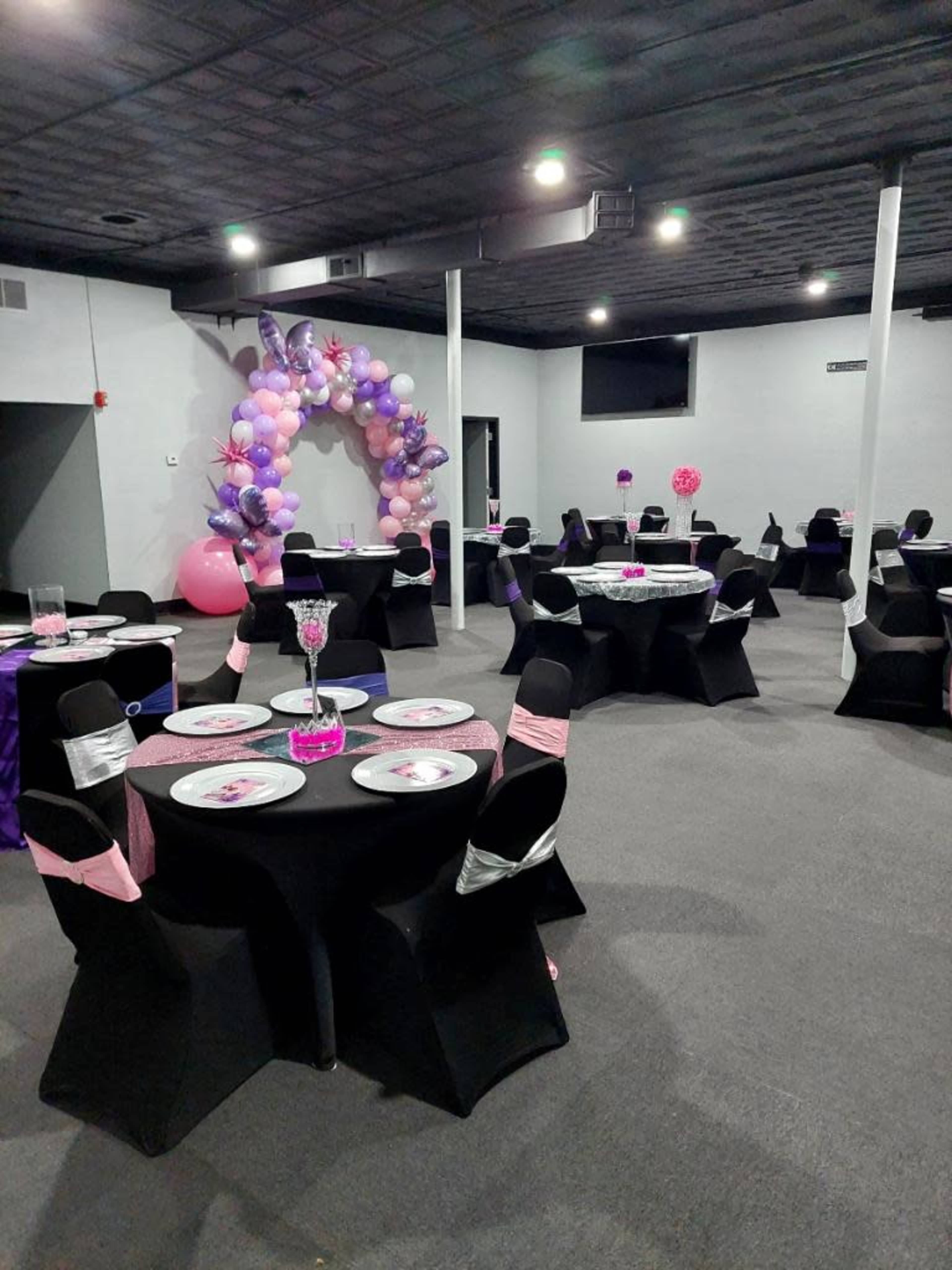 The image shows a party venue with black tables adorned with pink and purple decorations, surrounded by balloon arches and centerpieces.