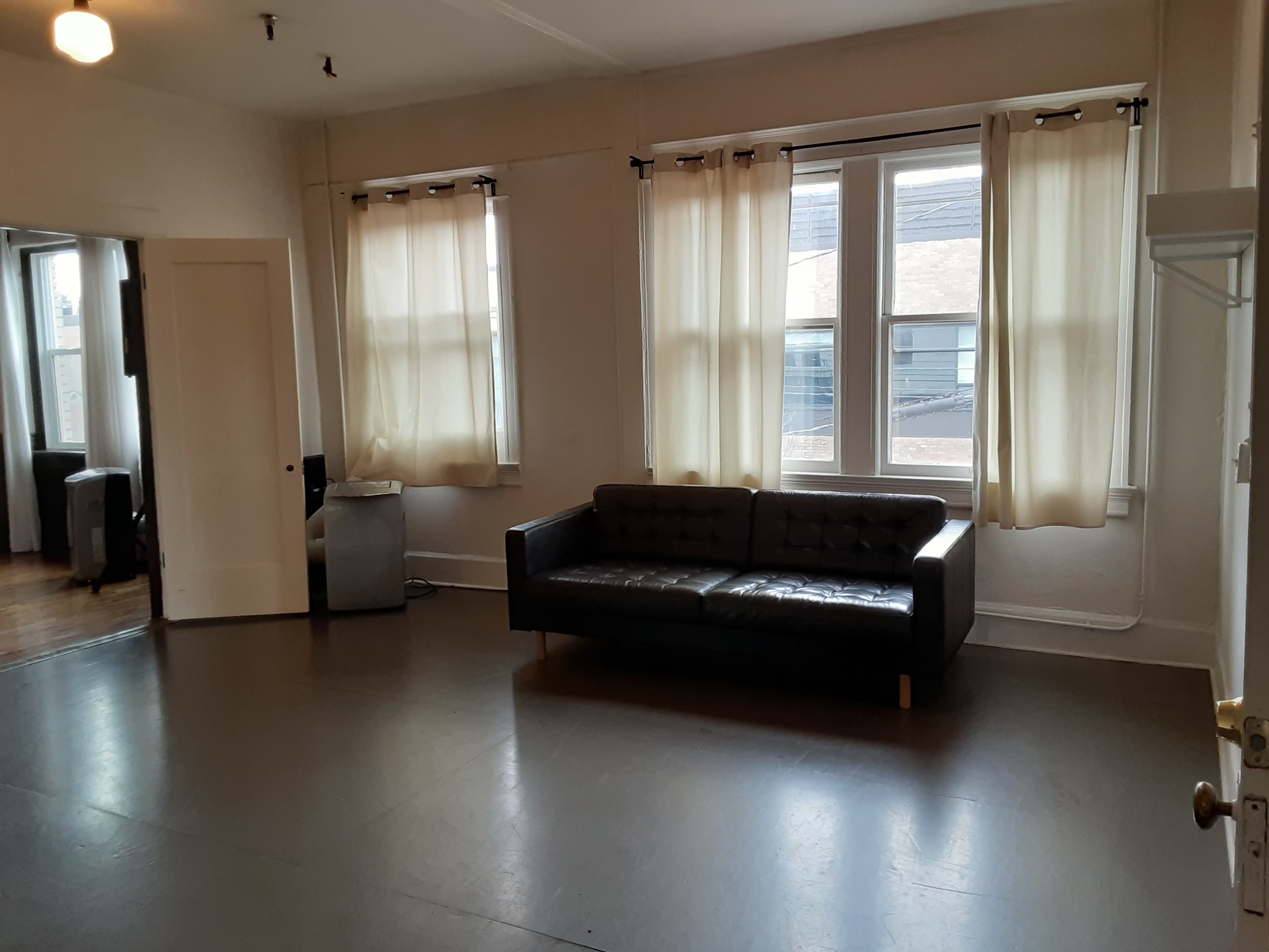 A spacious room with a black sofa, large windows with light curtains, and minimal furnishings.