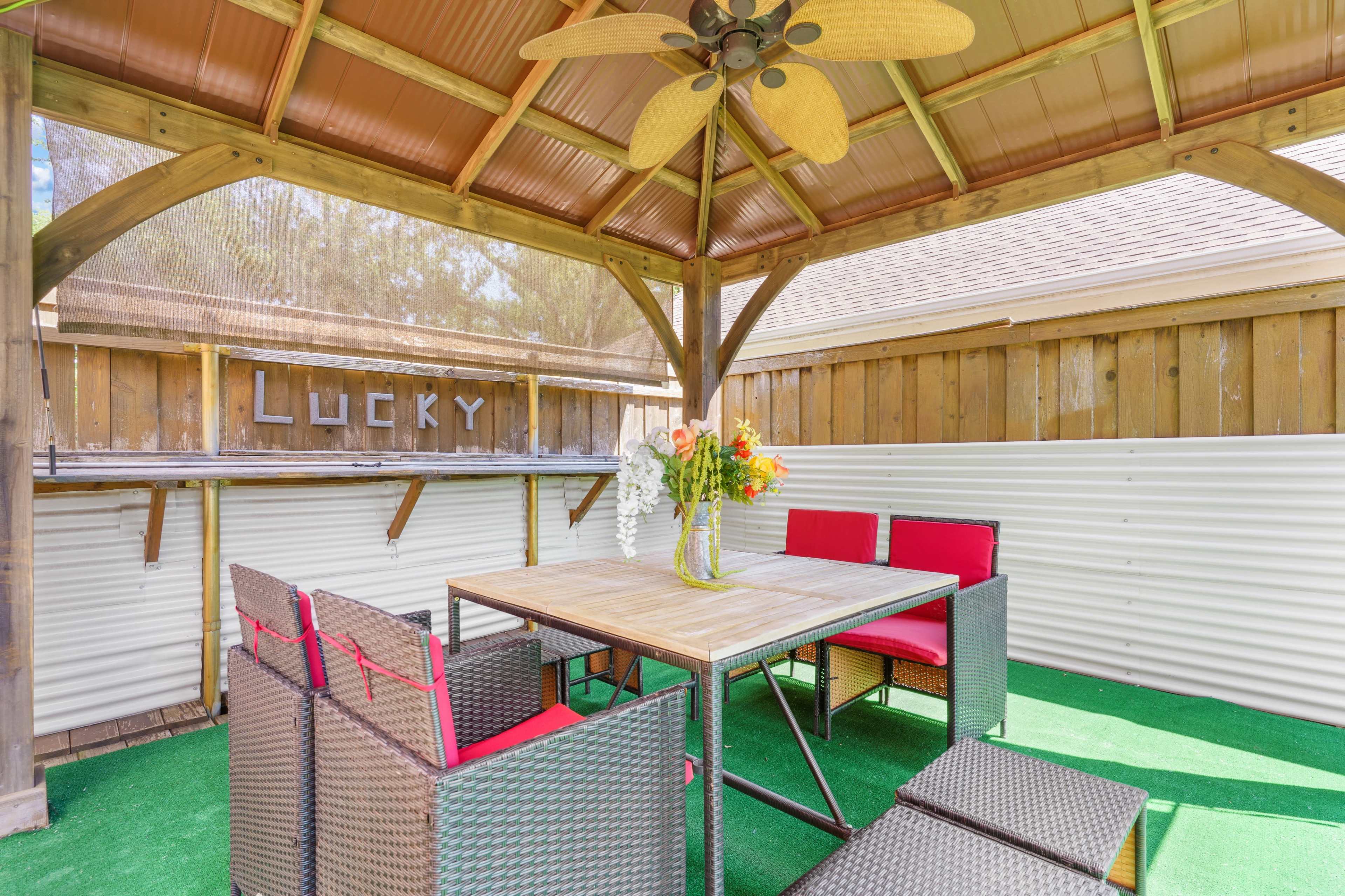 A shaded outdoor sitting area with a table, four red chairs, and a floral centerpiece under a wooden gazebo.