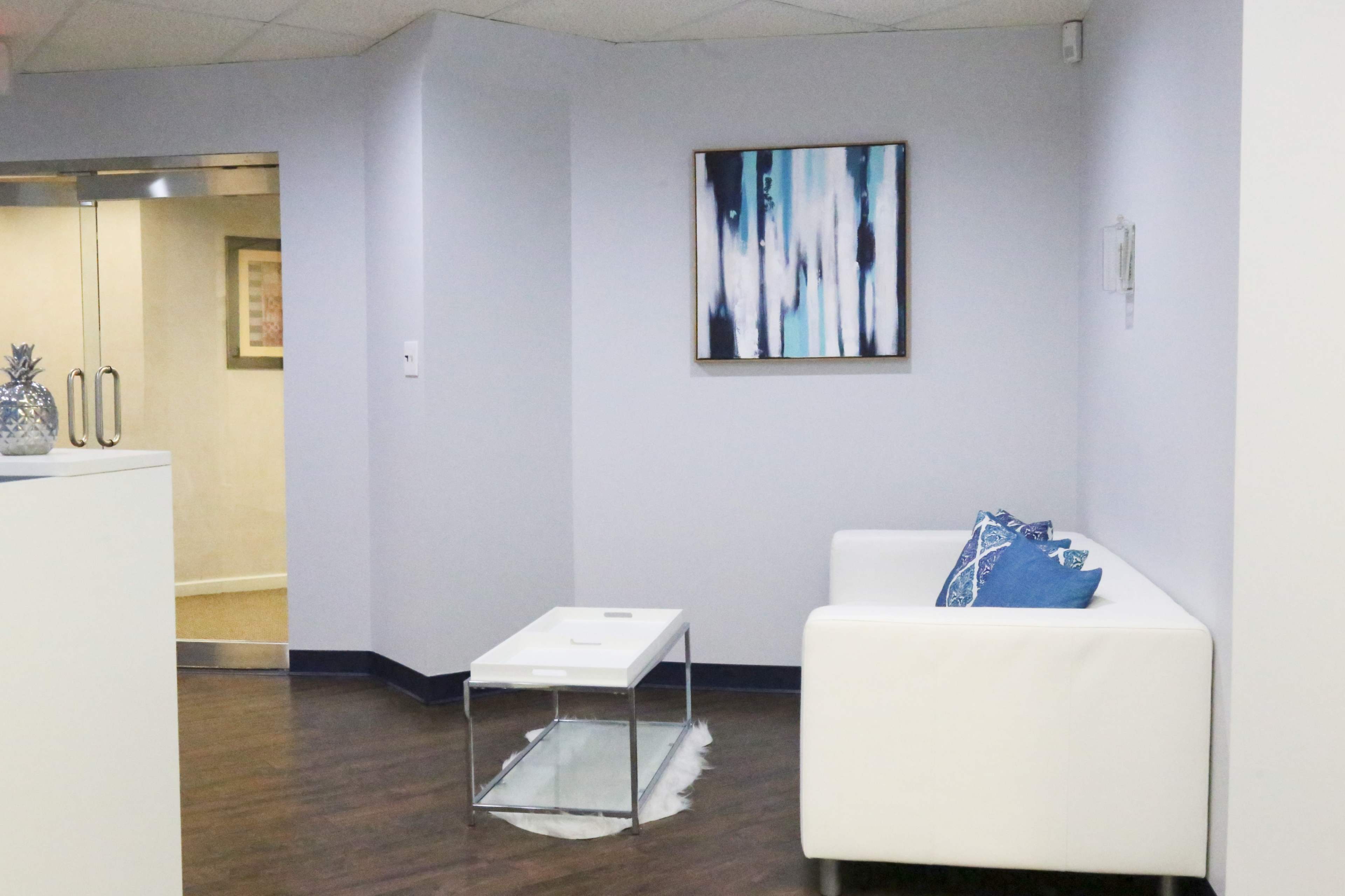 A modern waiting area with a white sofa, a glass coffee table, and a blue and white abstract painting on the wall.