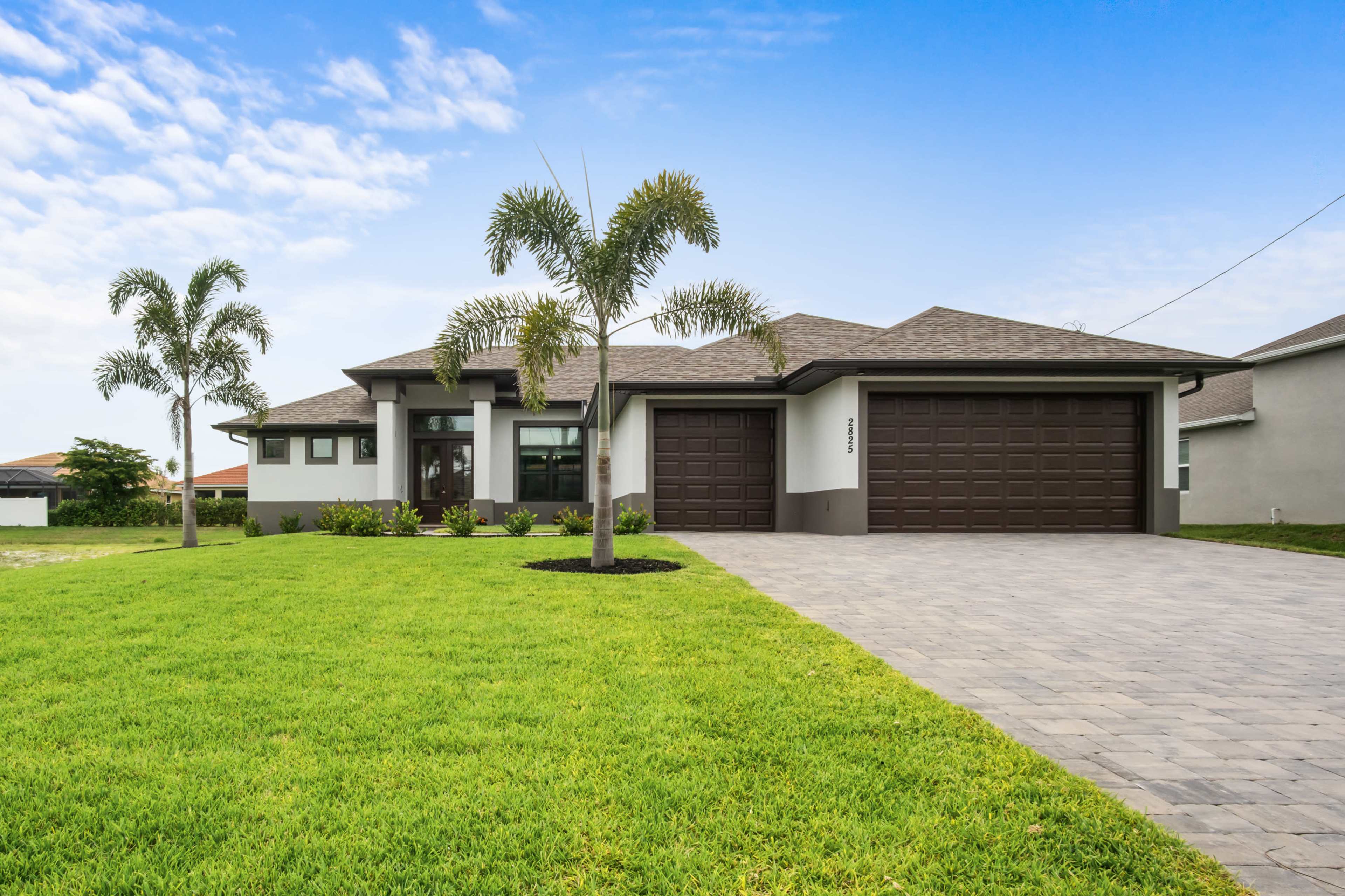 A modern single-story house with a two-car garage, surrounded by well-maintained grass and palm trees.